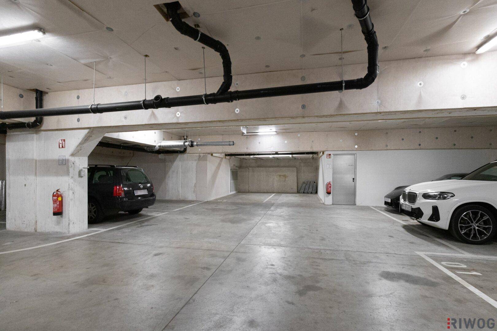 Geräumige Tiefgarage mit mehreren Stellplätzen, Betonwänden und sichtbaren Rohren an der Decke.