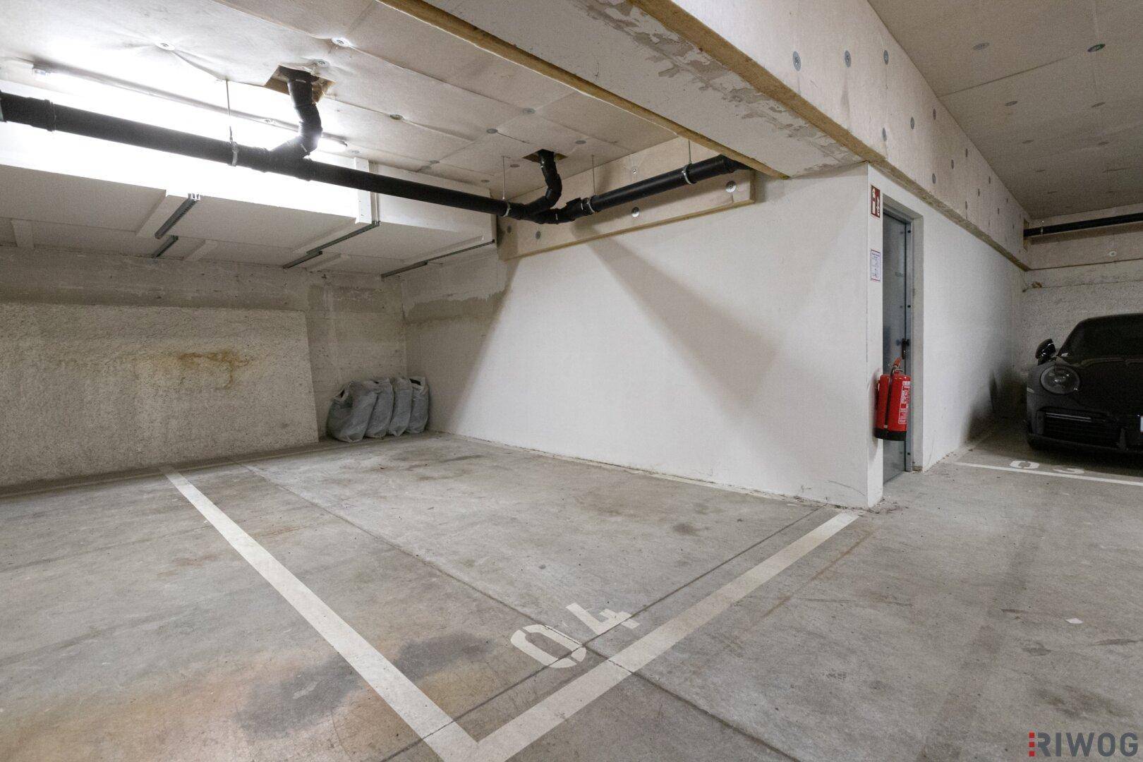 Leerer Tiefgaragenstellplatz mit Betonboden, Feuerlöscher und Zugang zu einem kleinen Aufzug.