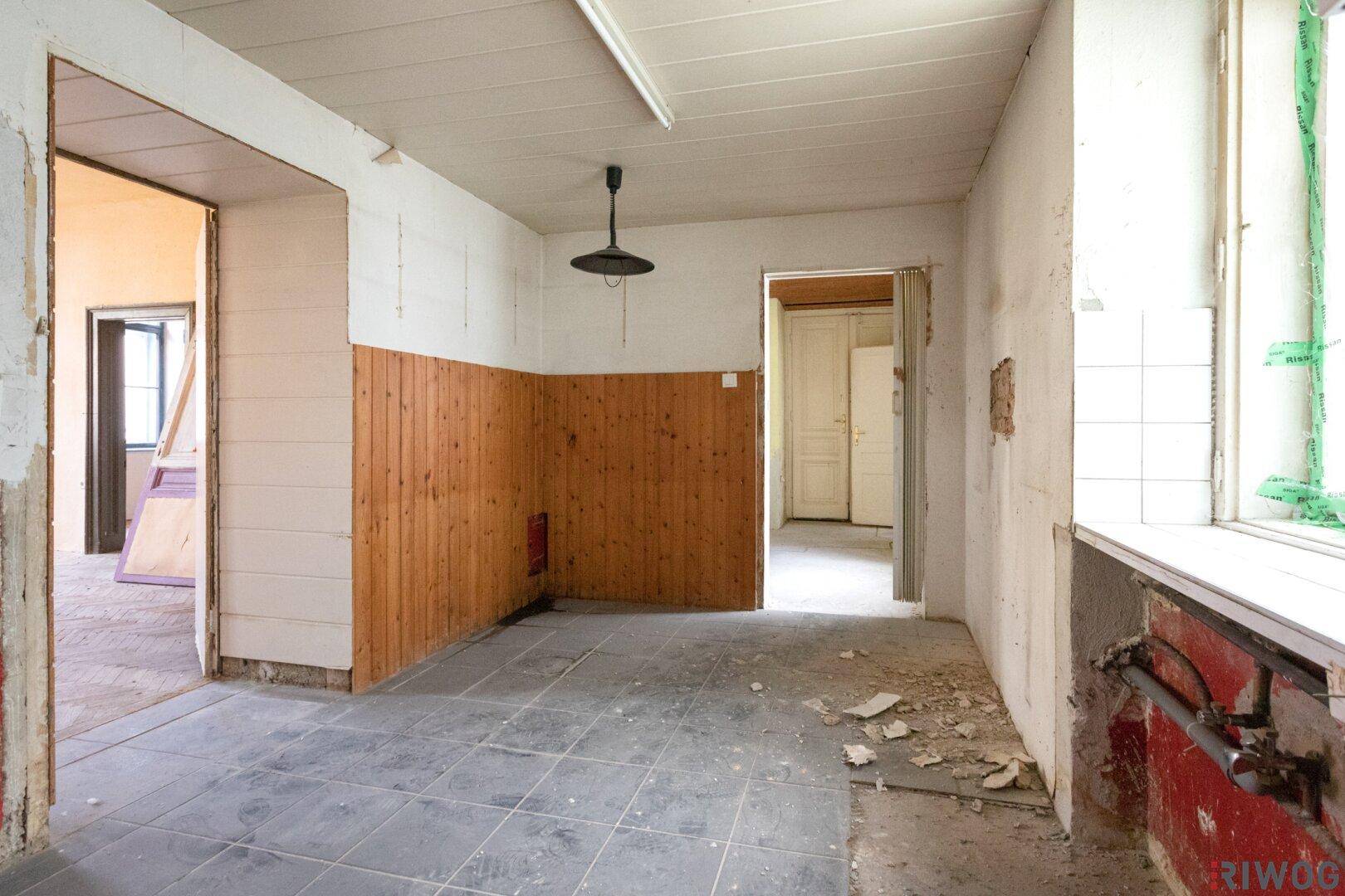 Unrenovierte Küche mit Fliesenboden, Holzverkleidung und Blick in einen weiteren Raum.