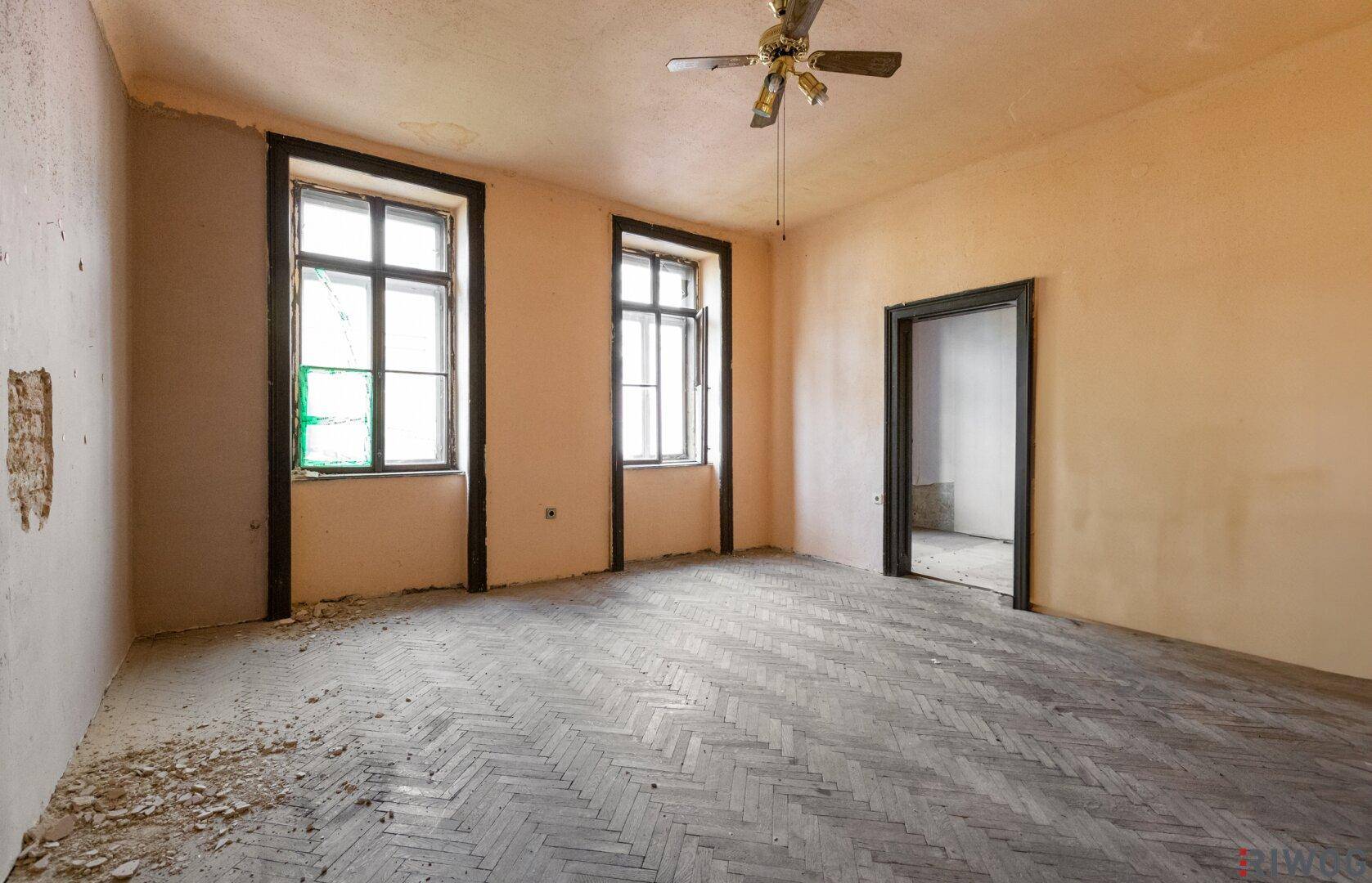 Unrenoviertes Zimmer mit Fischgrätparkett, hohen Decken und zwei Fenstern, bereit zur Sanierung.