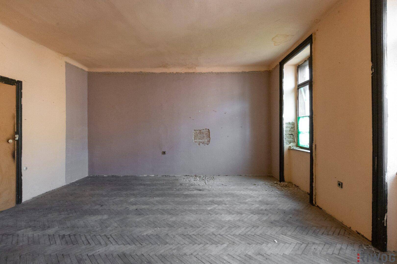 Leeres Zimmer mit abgenutztem Fischgrätparkett und teilweise gestrichenen Wänden, renovierungsbedürftig.