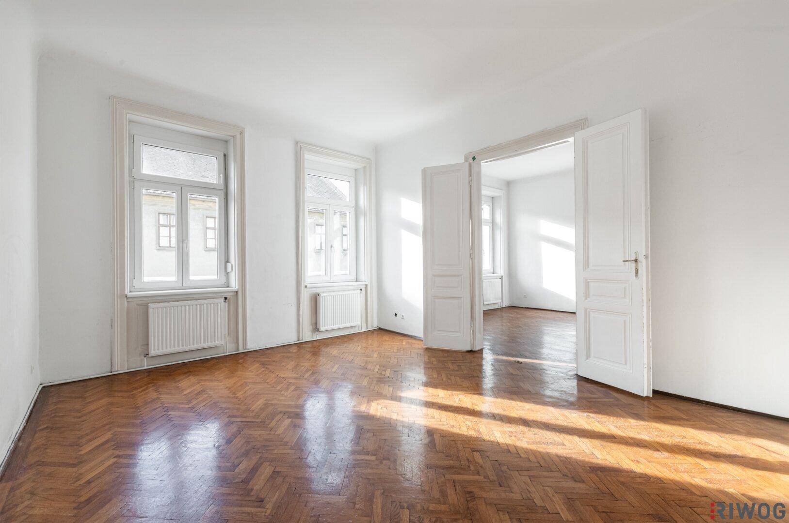 Geräumiges, leeres Wohnzimmer mit Fischgrätparkett und hohen Fenstern, bereit zur Einrichtung.