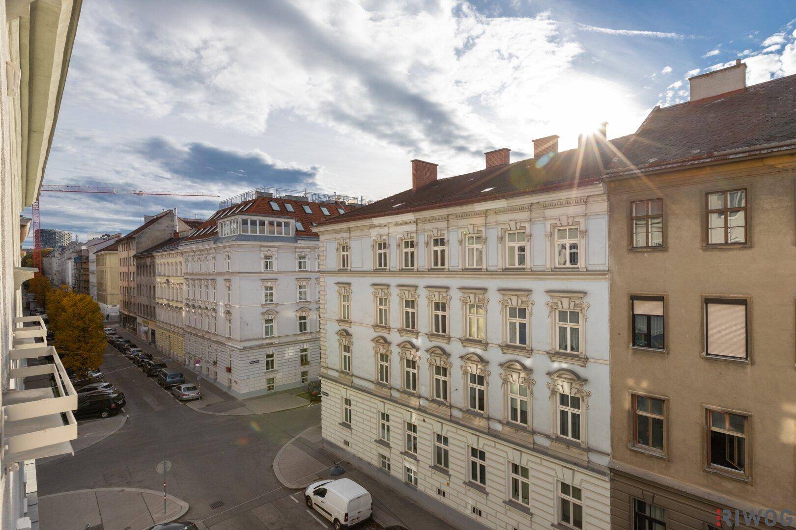 Blick vom Balkon auf eine belebte Stadtstraße mit historischen Gebäuden und sonnigem Himmel.