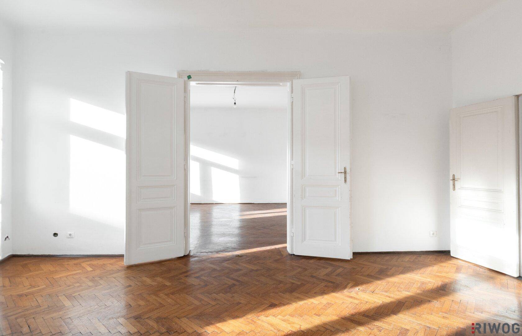 Leeres Zimmer mit Fischgrätparkett und doppelten Flügeltüren, die zu einem weiteren Raum führen.