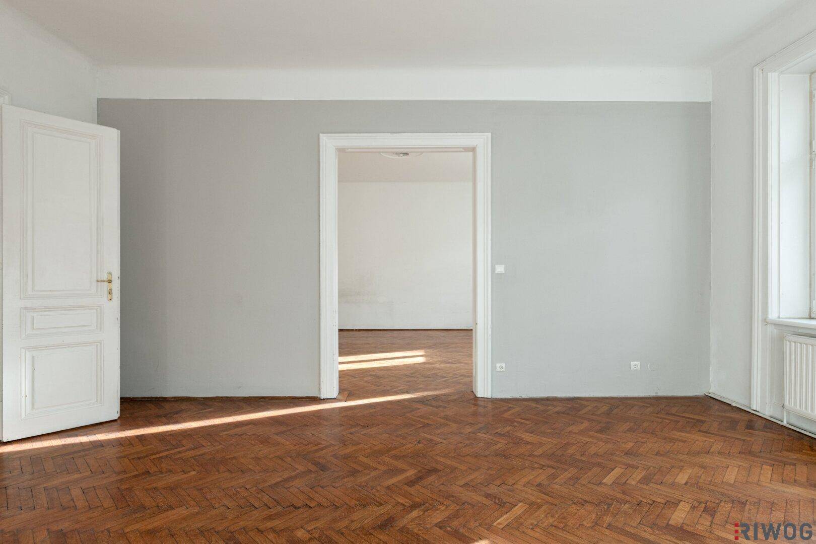 Geräumiges Zimmer mit Fischgrätparkett und einer grauen Wand, die zu einem weiteren Raum führt.