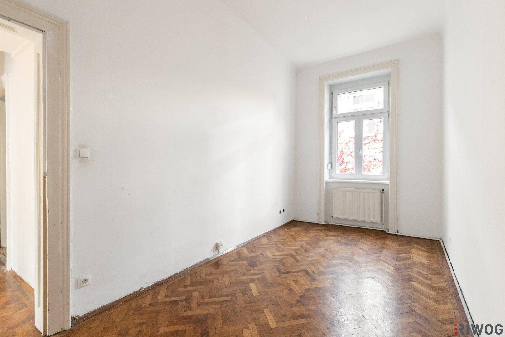 Helles, leeres Schlafzimmer mit Fischgrätparkett und einem großen Fenster, bereit zur Einrichtung.