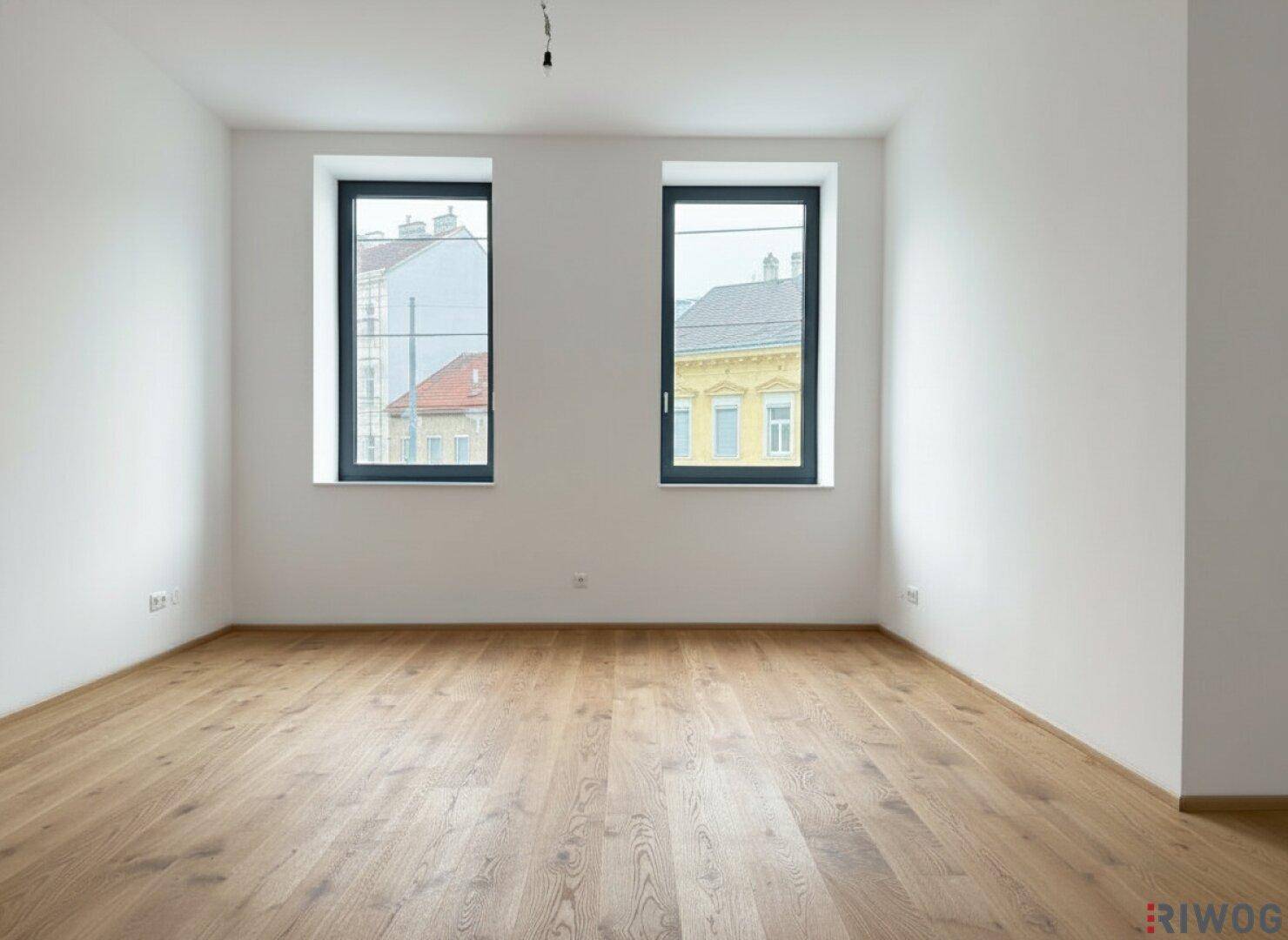 Helles Zimmer mit neuem Holzparkettboden und zwei großen Fenstern, die einen Blick auf die Stadt bieten.