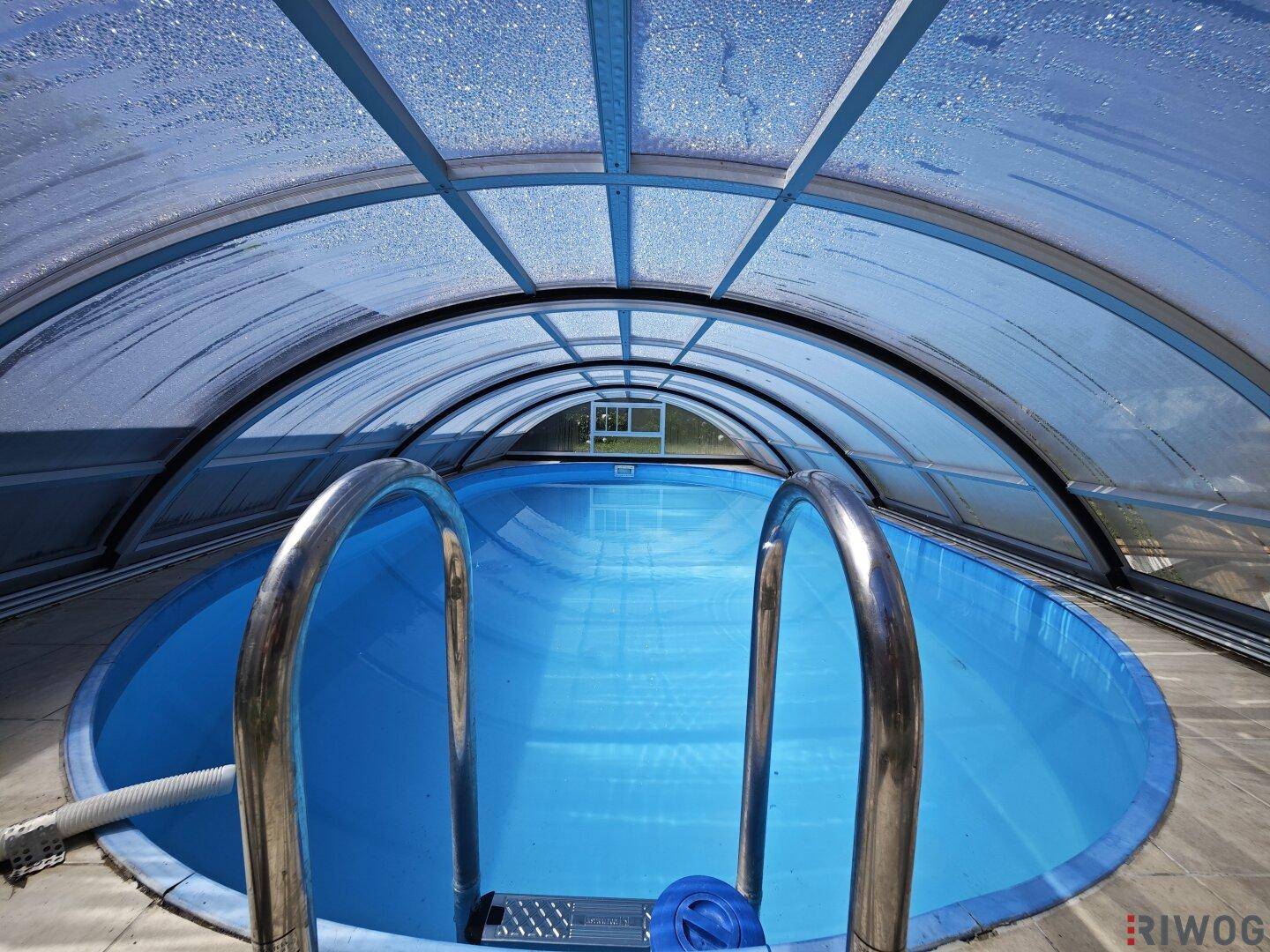 Überdachter Swimmingpool mit blauer Auskleidung und Metallleiter unter einem transparenten Dach.