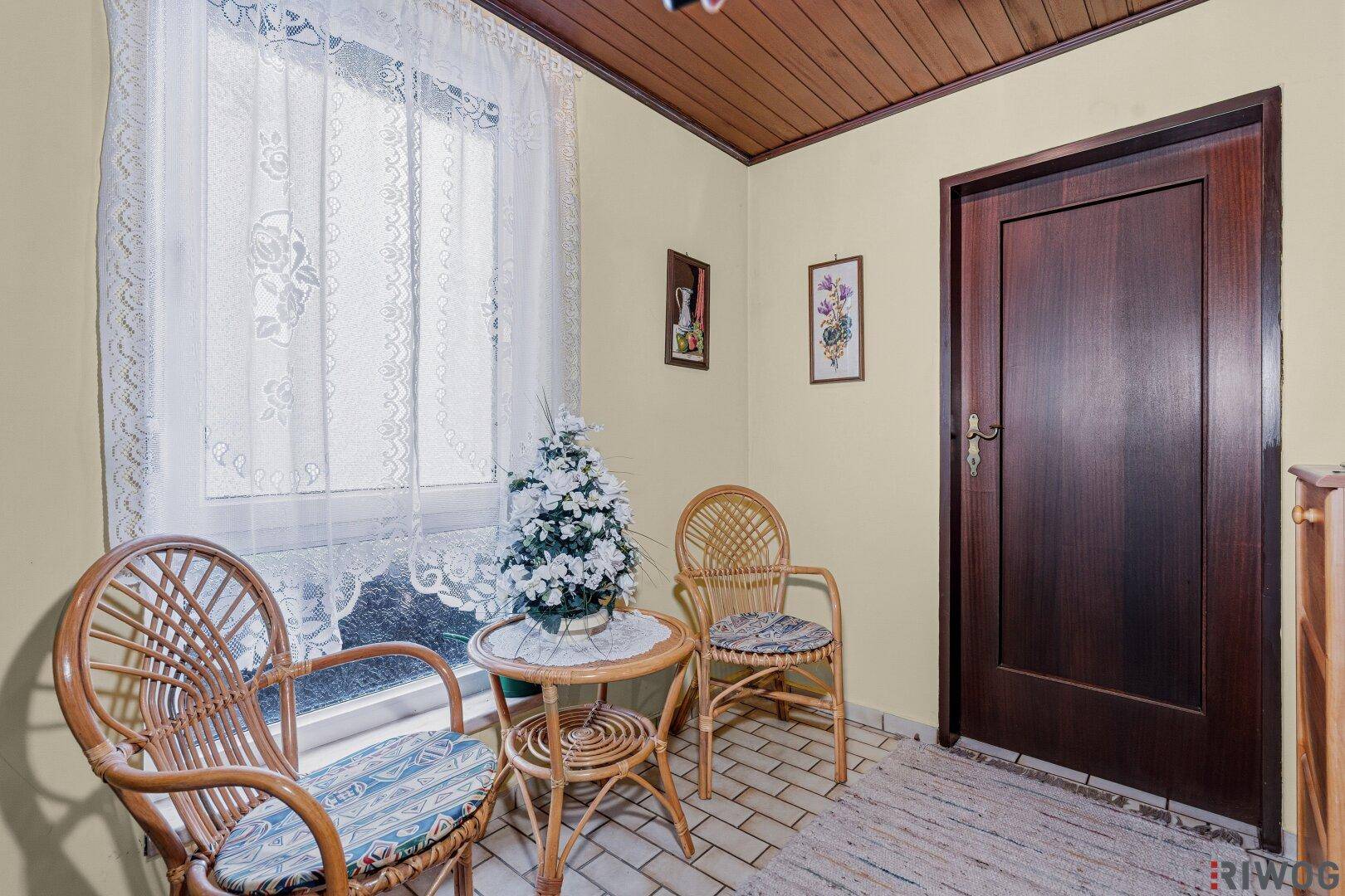 Gemütlicher Eingangsbereich mit Korbmöbeln, einem Fenster mit Spitzenvorhängen und Fliesenboden.