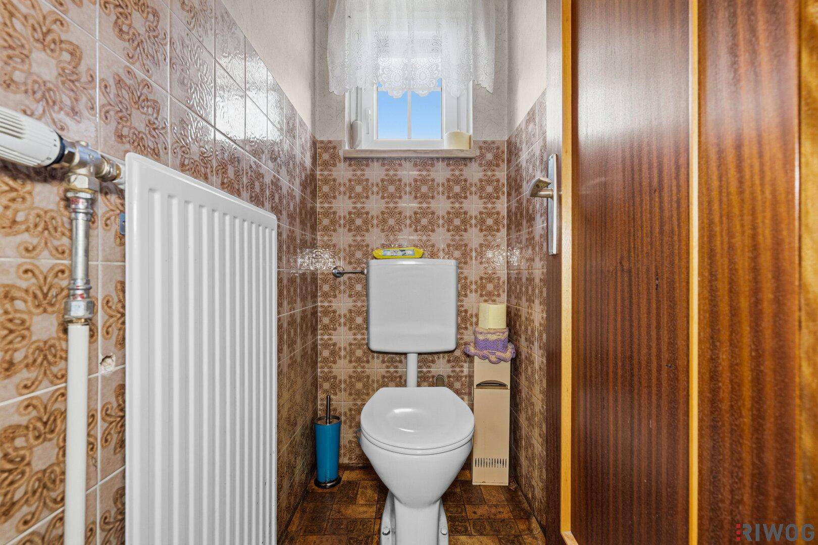Kleines Gäste-WC mit Toilette, Heizkörper und braunen Fliesen, ausgestattet mit einem Fenster.