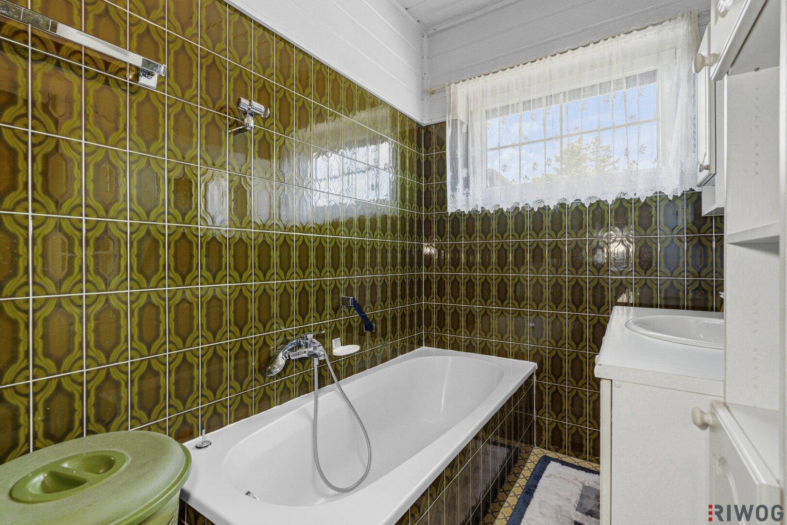 Badezimmer mit Badewanne, Waschbecken und auffälligen grün-braunen Fliesen, inklusive Fenster.