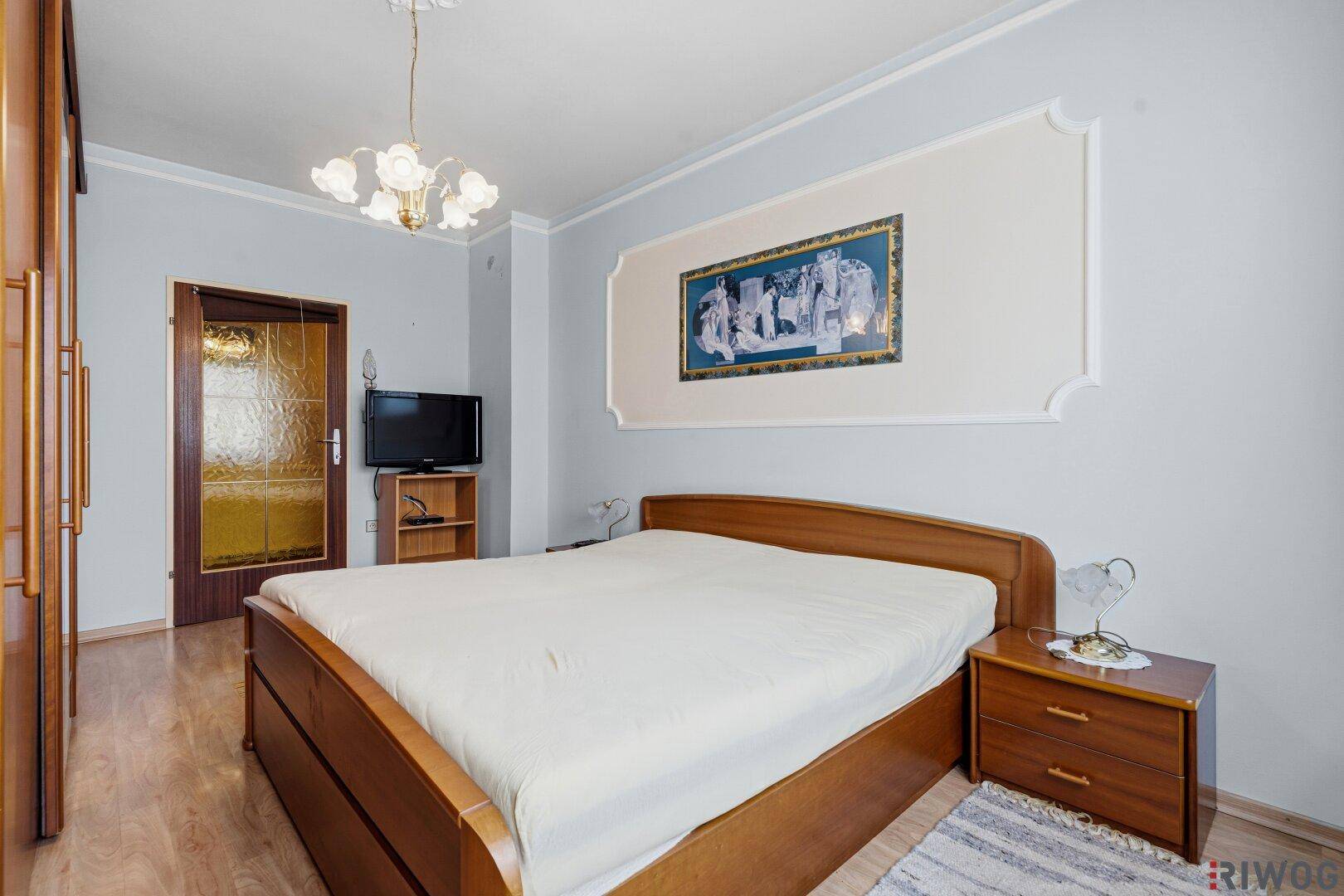 Schlafzimmer mit Holzdoppelbett, Nachttischen, Kleiderschrank und hellblauen Wänden mit Stuckverzierung.