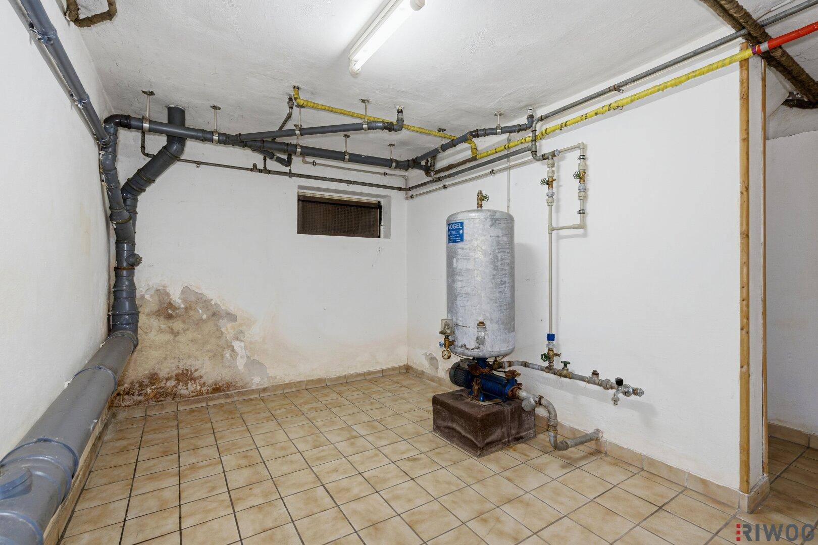 Hauswirtschaftsraum im Keller mit sichtbaren Rohren, Warmwasserbereiter und Feuchtigkeitsspuren an der Wand.