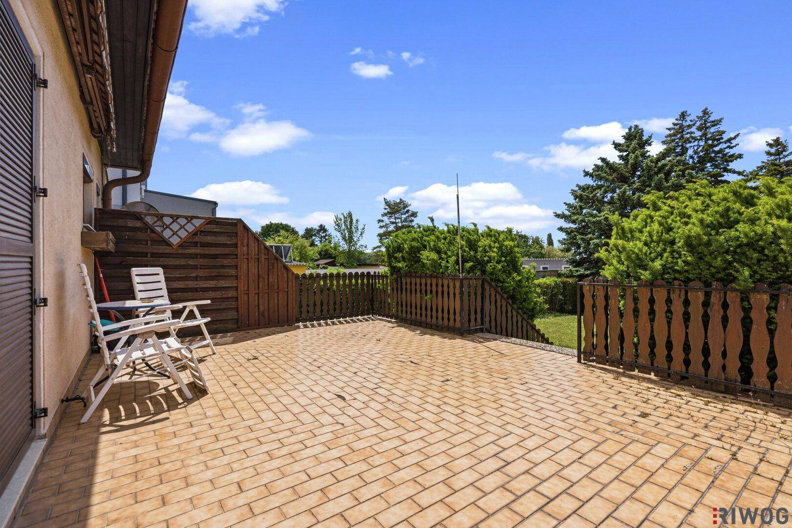 Große gepflasterte Terrasse mit Gartenmöbeln, Holzzaun und grünen Bäumen im Hintergrund.
