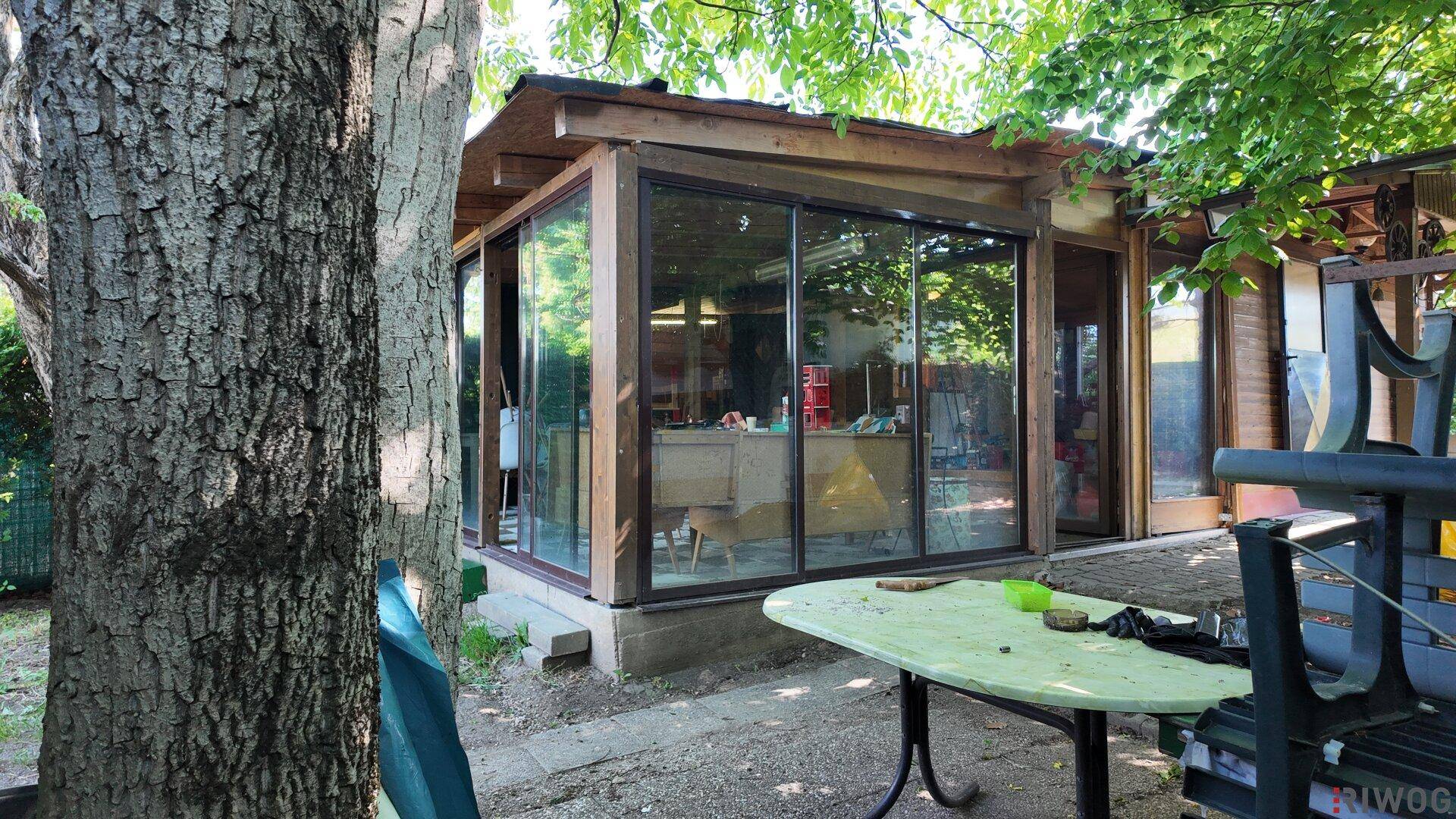 Wintergarten mit großen Glasfronten und einem Tisch im Freien unter einem Baum.