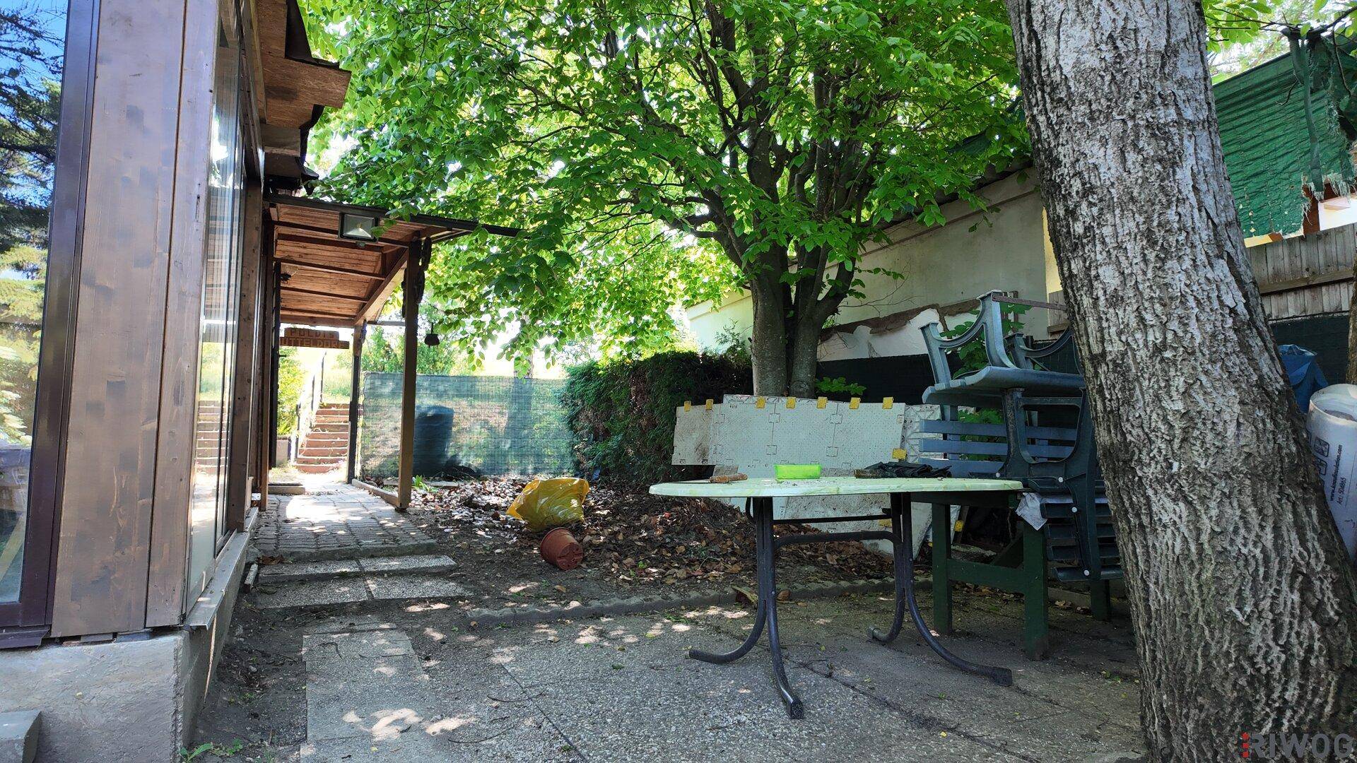 Gartenbereich mit Bäumen, einer kleinen Terrasse und Gartenmöbeln, umgeben von viel Grün.