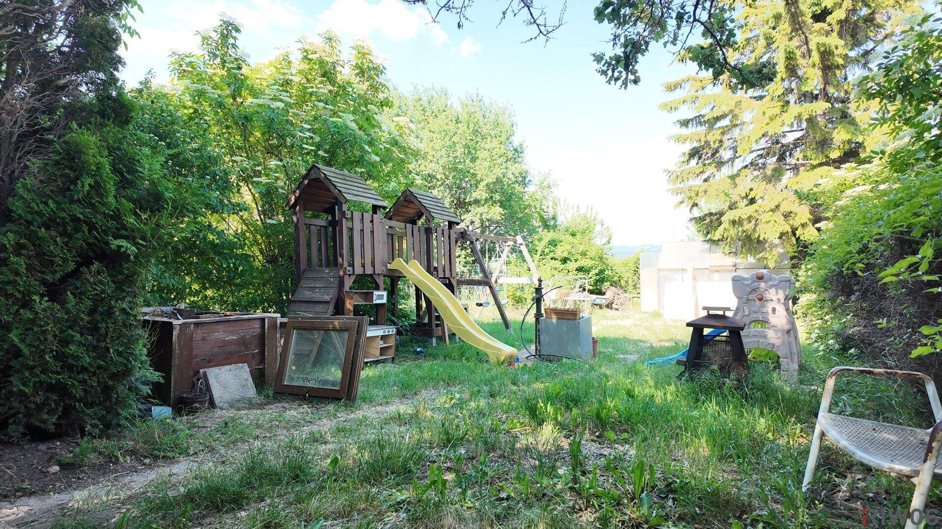Großer Garten mit Spielplatz, Rutsche und Schaukeln, umgeben von Bäumen und viel Grün.