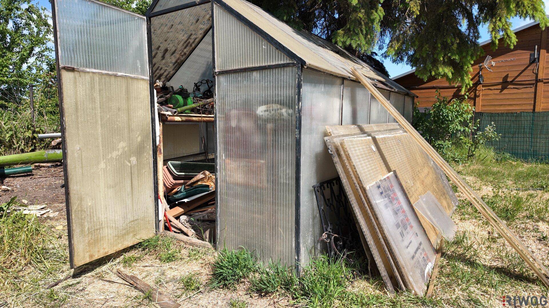 Gewächshaus mit offener Tür und gelagerten Materialien im Gartenbereich.
