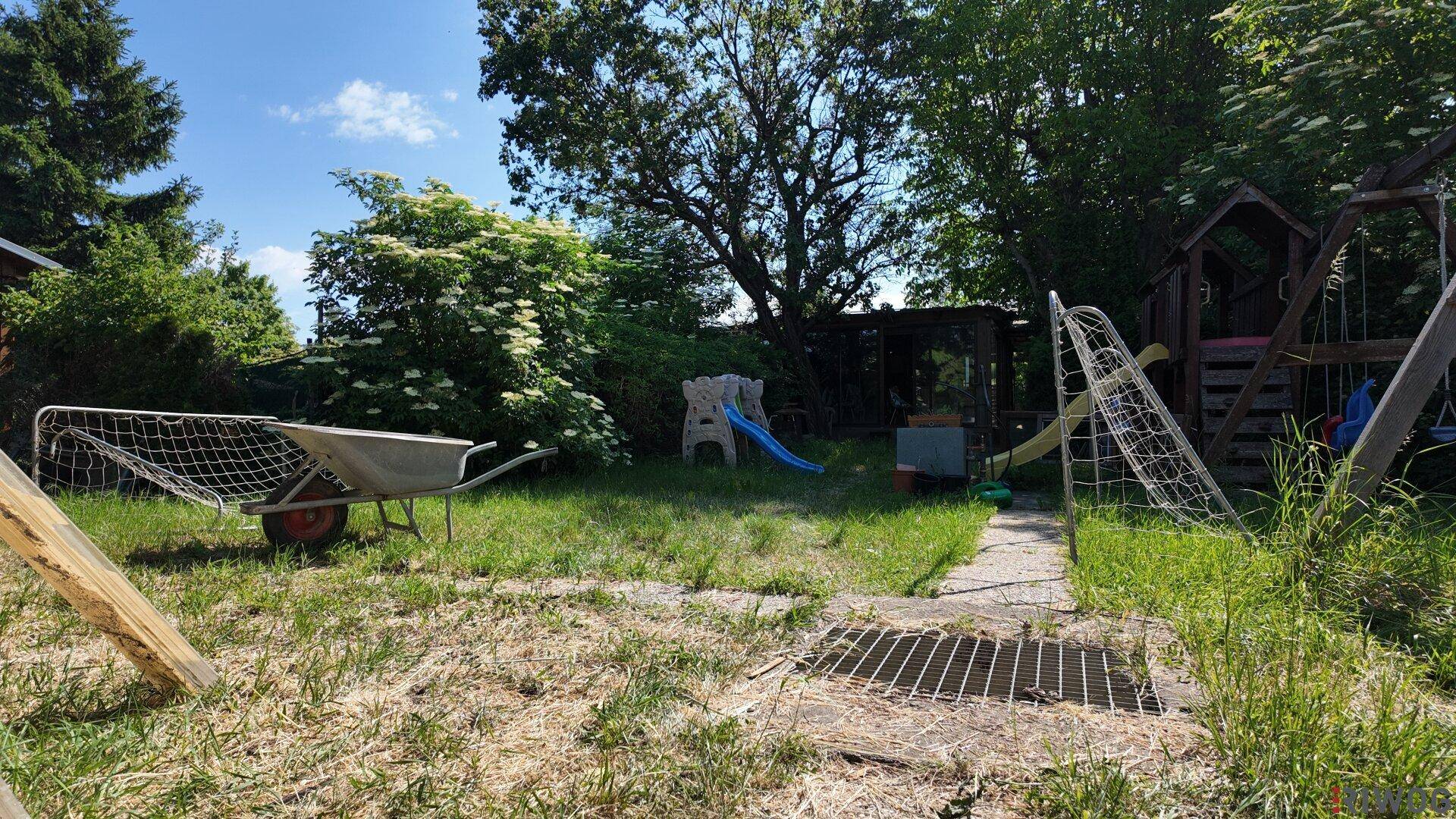 Sonniger Gartenbereich mit Rasen, einem Schubkarren und einem Spielplatz im Hintergrund.