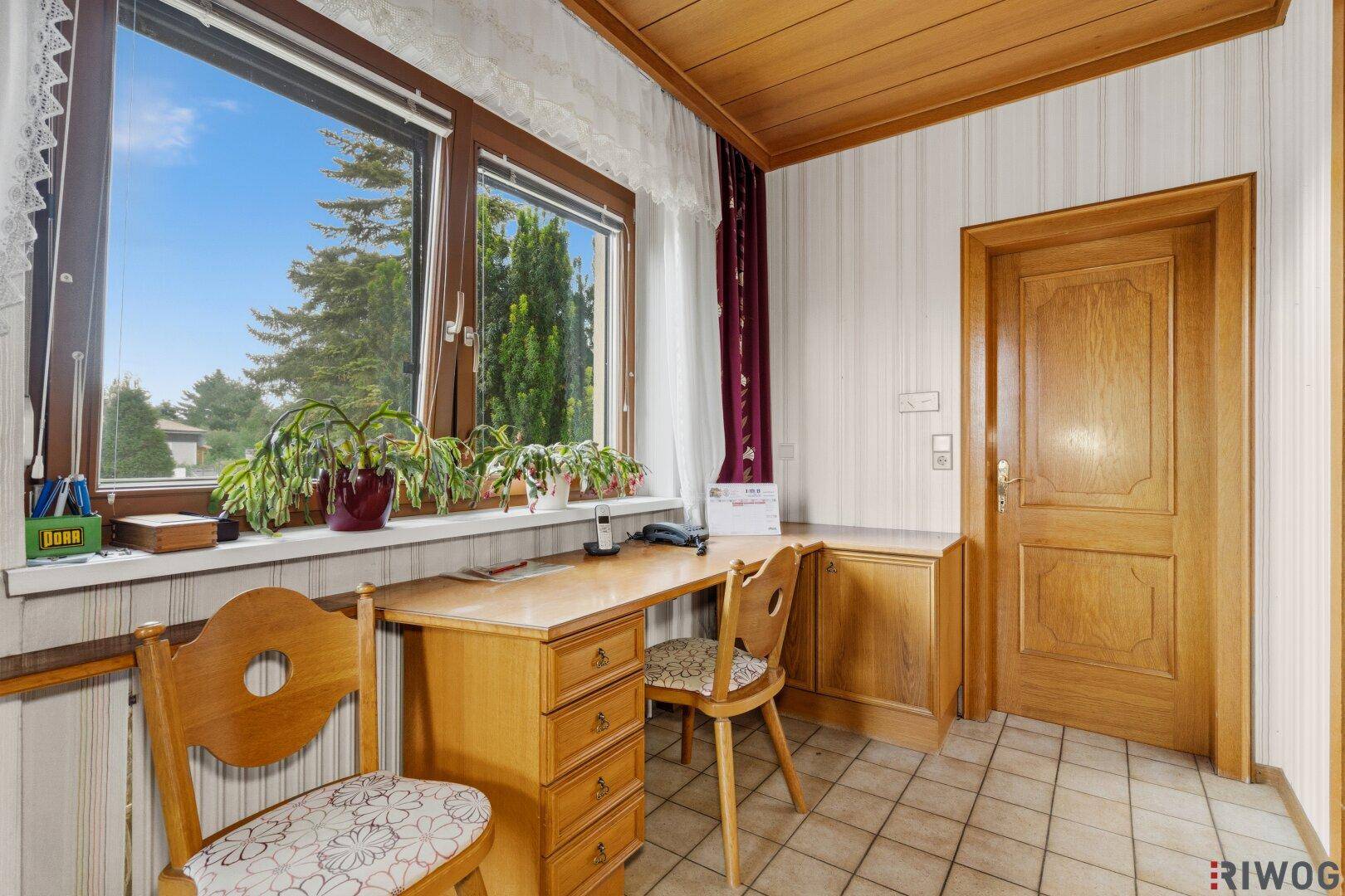Helles Arbeitszimmer mit Holzdecke, gefliestem Boden und Blick ins Grüne durch große Fenster.