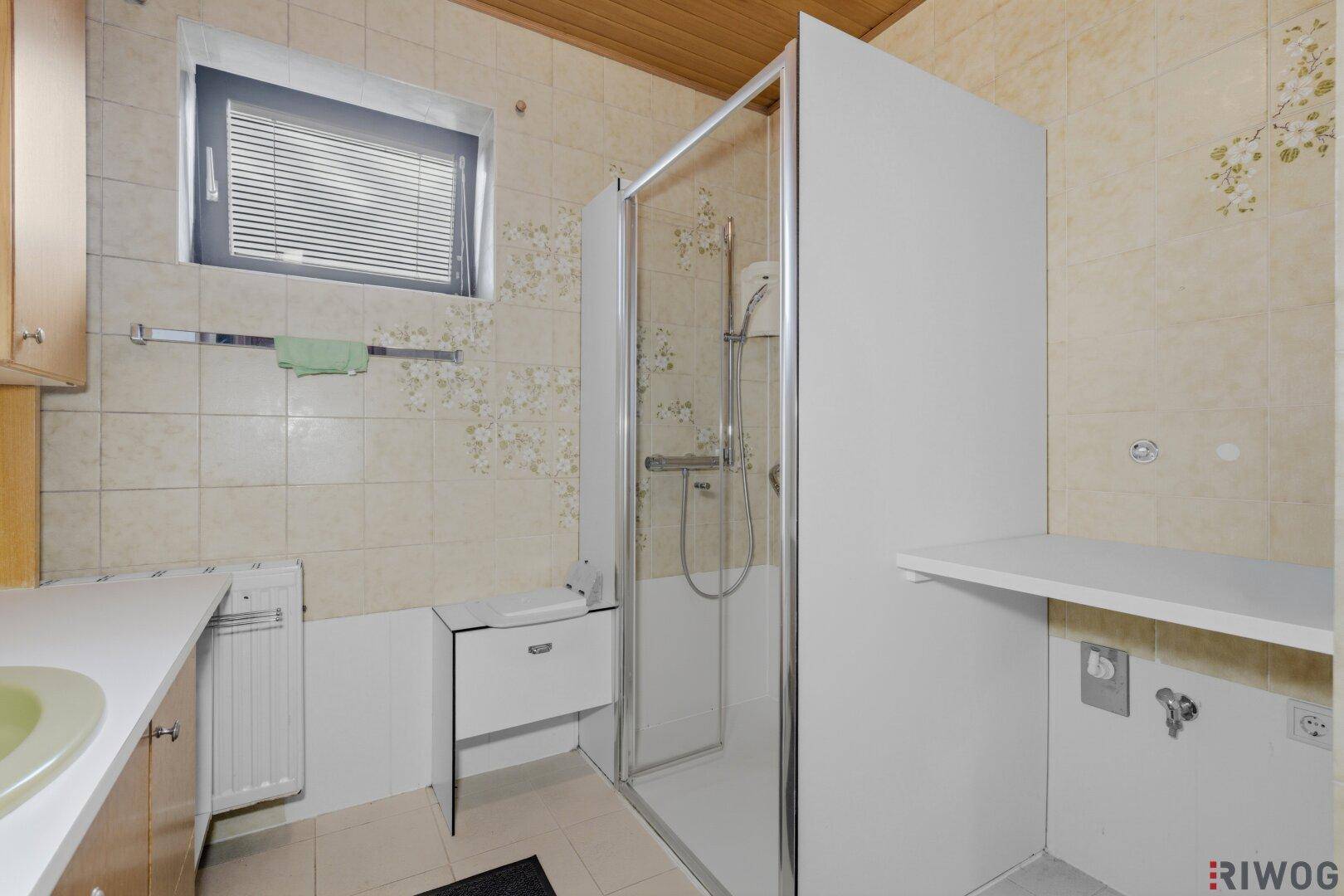 Duschbereich im Badezimmer mit beigen Fliesen, Duschkabine und Fenster für Tageslicht.