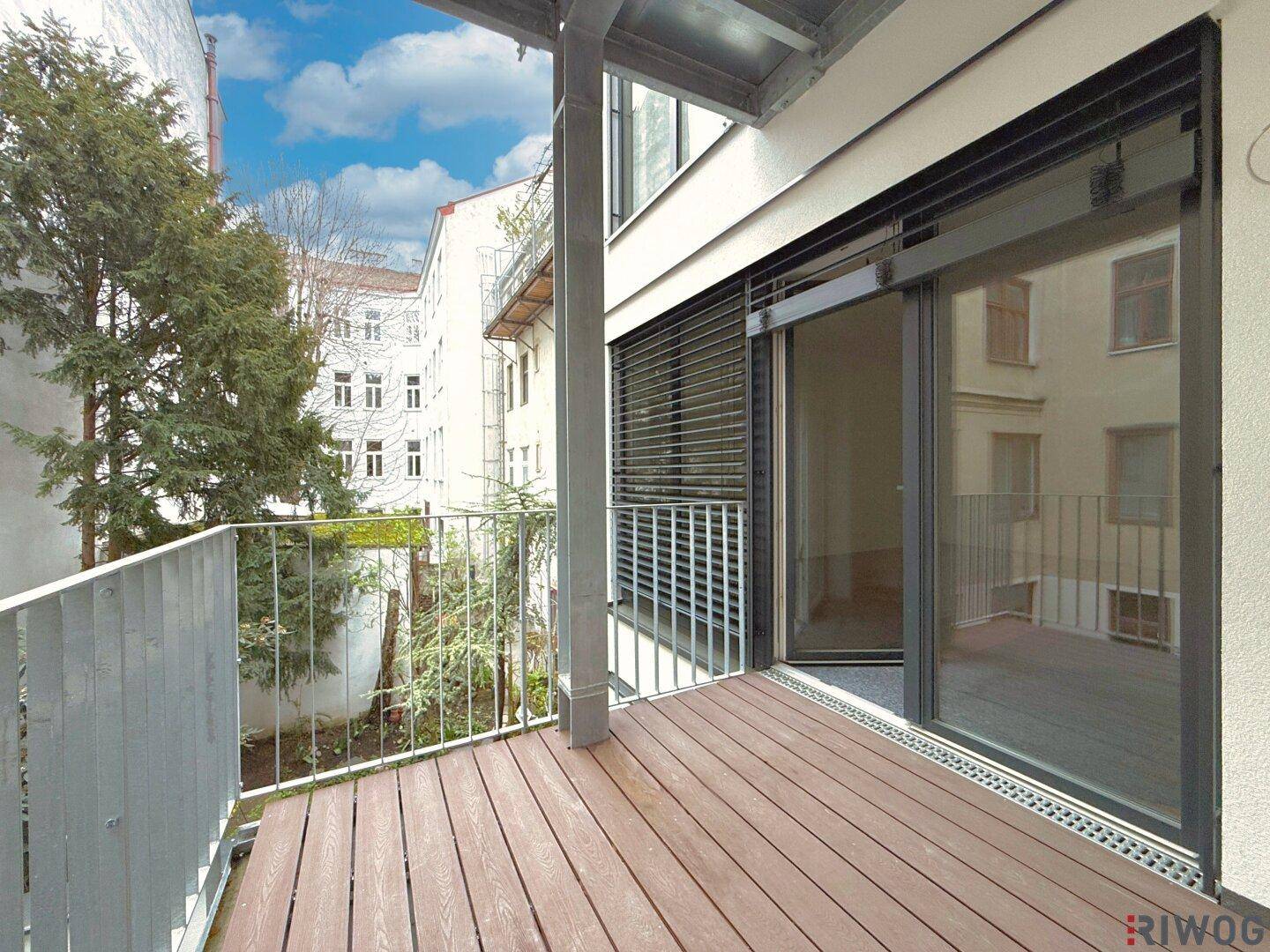 Geräumiger Balkon mit Holzdielenboden, modernem Metallgeländer und Schiebetür, bietet Blick auf umliegende Gebäude und Bäume.