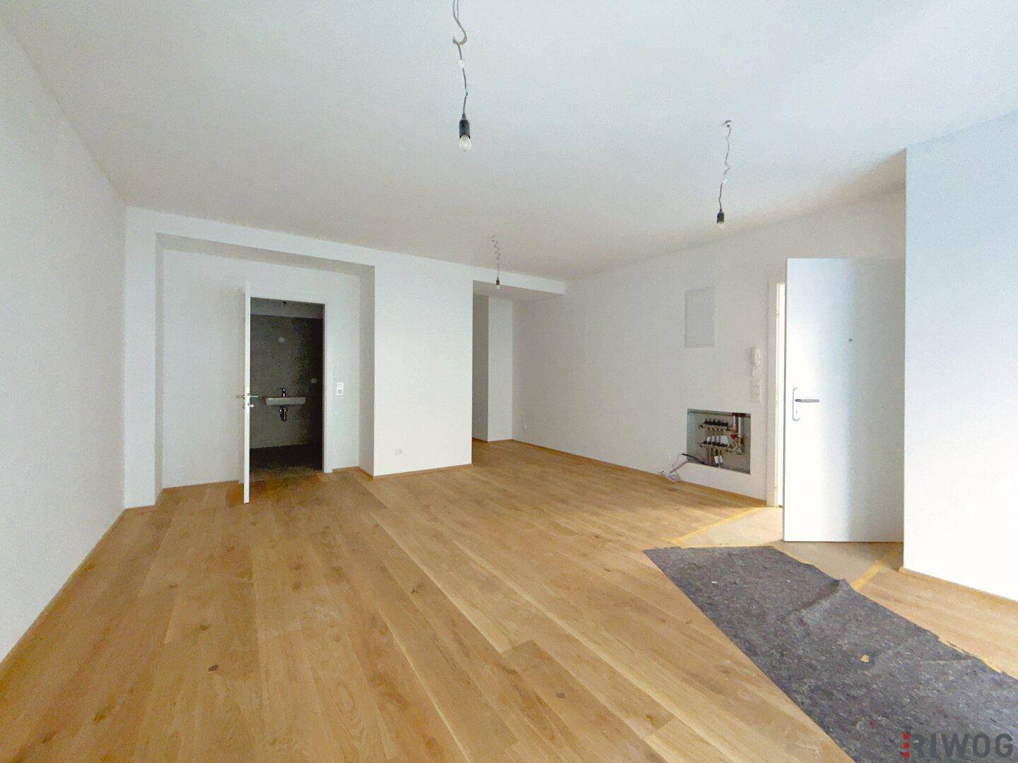 Geräumiger, unmöblierter Raum mit hellem Holzfußboden und Zugang zu einem Badezimmer im Rohbau.