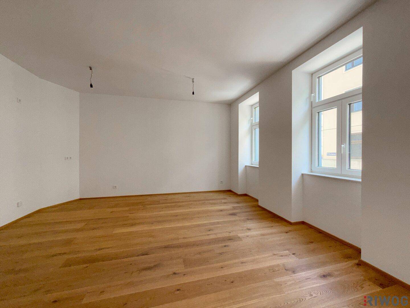 Geräumiger, heller Wohnbereich mit elegantem Holzboden und zwei Fenstern, ideal für Wohnzwecke.