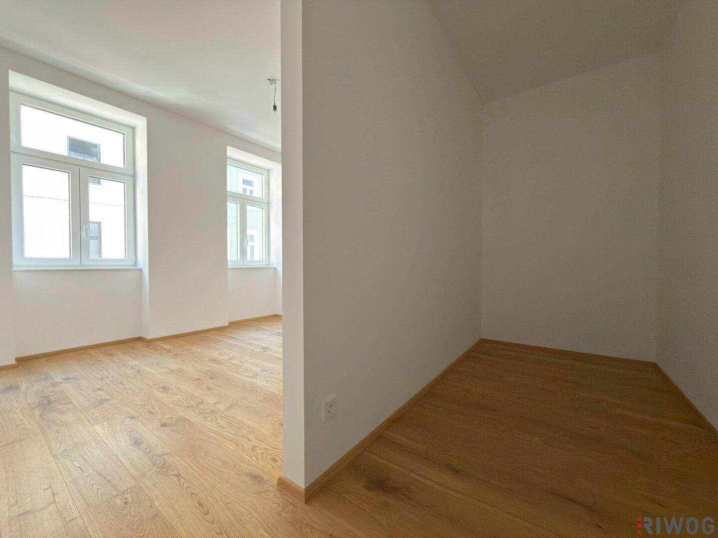 Heller Raum mit Holzboden und zwei Fenstern, bietet Potenzial für verschiedene Nutzungen und Stadtblick.