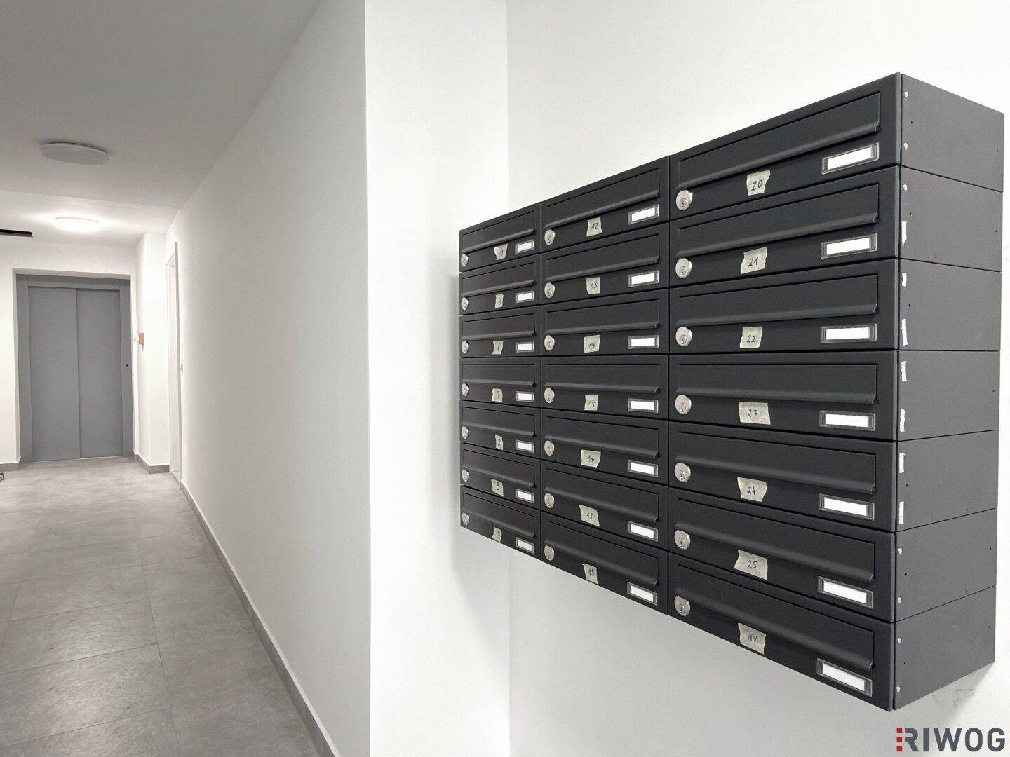 Langer Flur mit Wandbriefkästen und einem Aufzug im Hintergrund, alles in hellem Design.