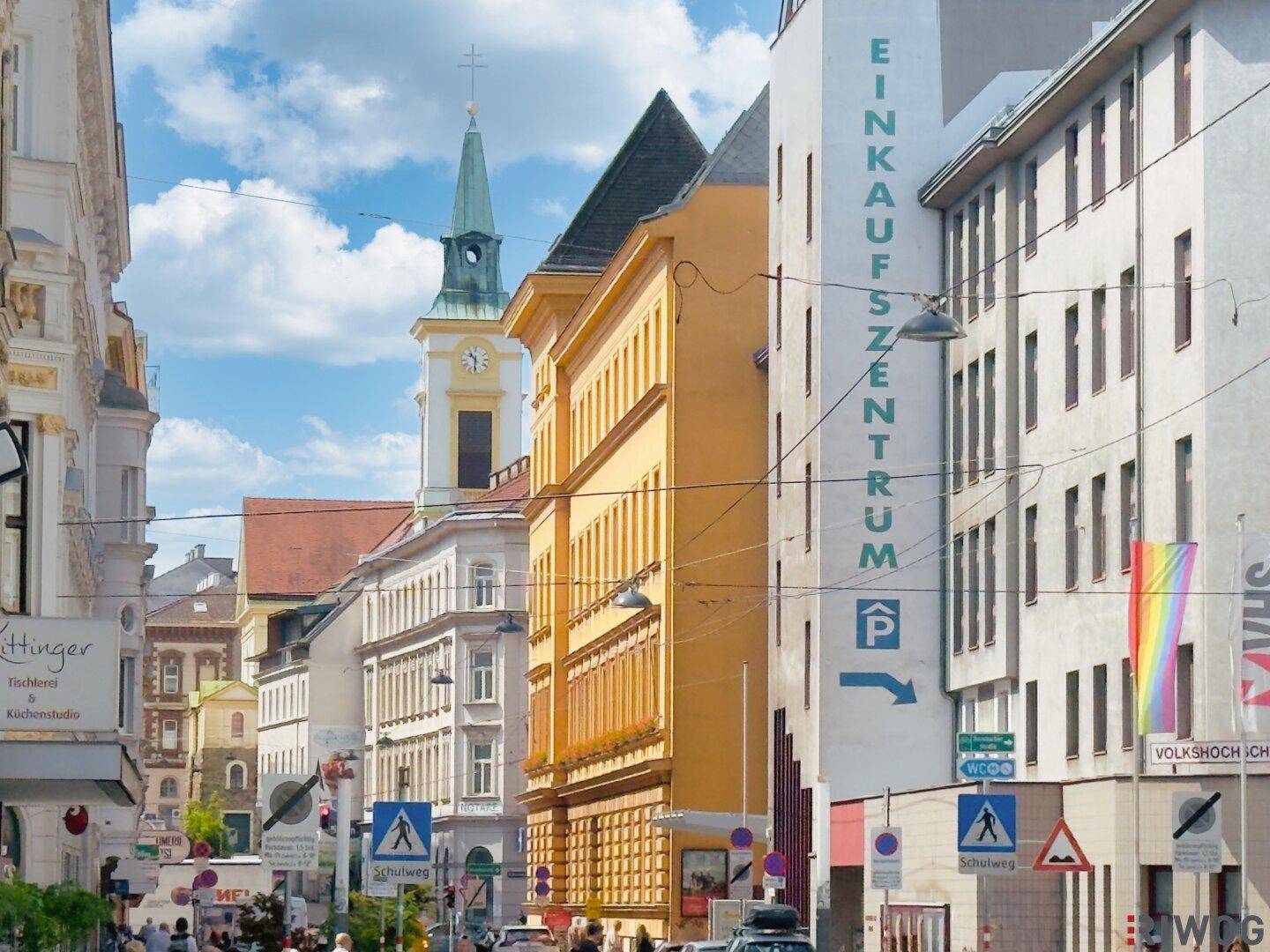 Belebte Stadtstraße mit historischen Gebäuden, einem Kirchturm und einem Einkaufzentrumsschild.