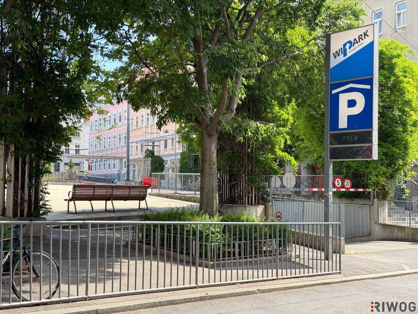 Einfahrt zu einer Tiefgarage mit Parkplatzschild, umgeben von Bäumen und einer Sitzbank.