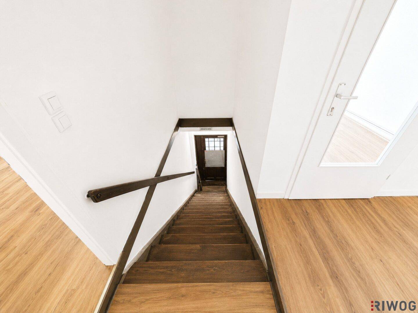 Holztreppe führt nach unten, umgeben von weißen Wänden und einem Laminatboden im oberen Bereich.