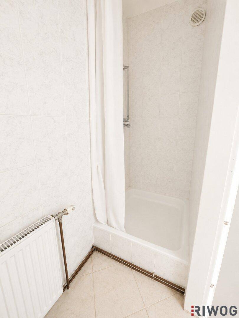 Kleines Badezimmer mit Duschkabine, weißen Fliesen an Wänden und Boden sowie einem Heizkörper.