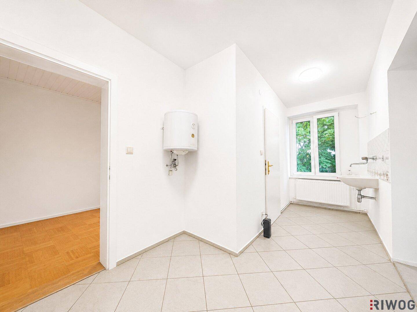 Funktionaler Raum mit Fliesenboden, Waschbecken und Warmwasserboiler, angrenzend an einen Raum mit Parkett.