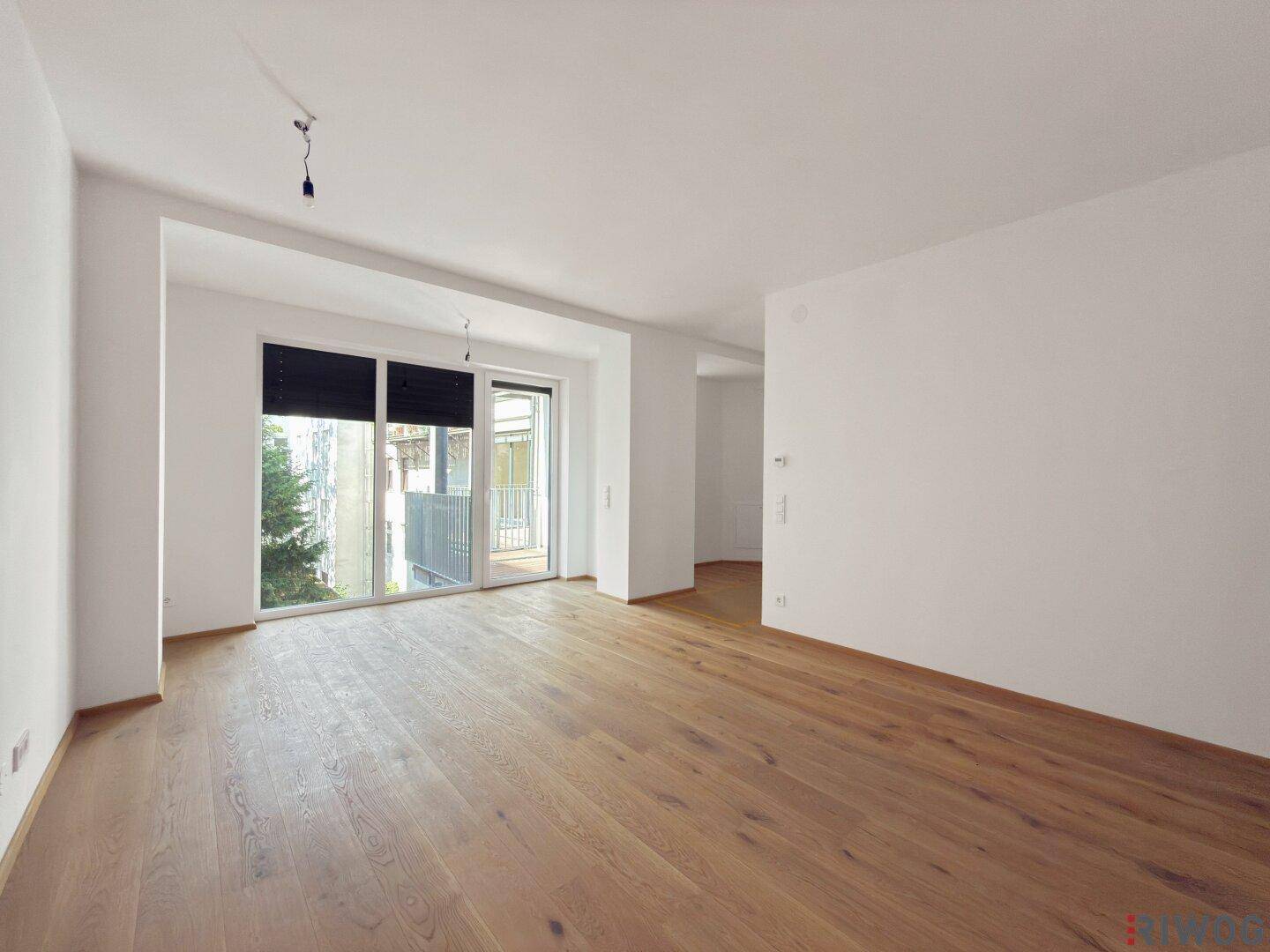 Helles, leeres Wohnzimmer mit neuem Parkettboden und Zugang zu einem Balkon.