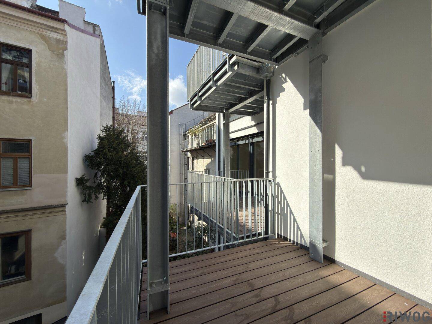 Neuer Balkon mit Holzboden und Metallgeländer, umgeben von Gebäuden.
