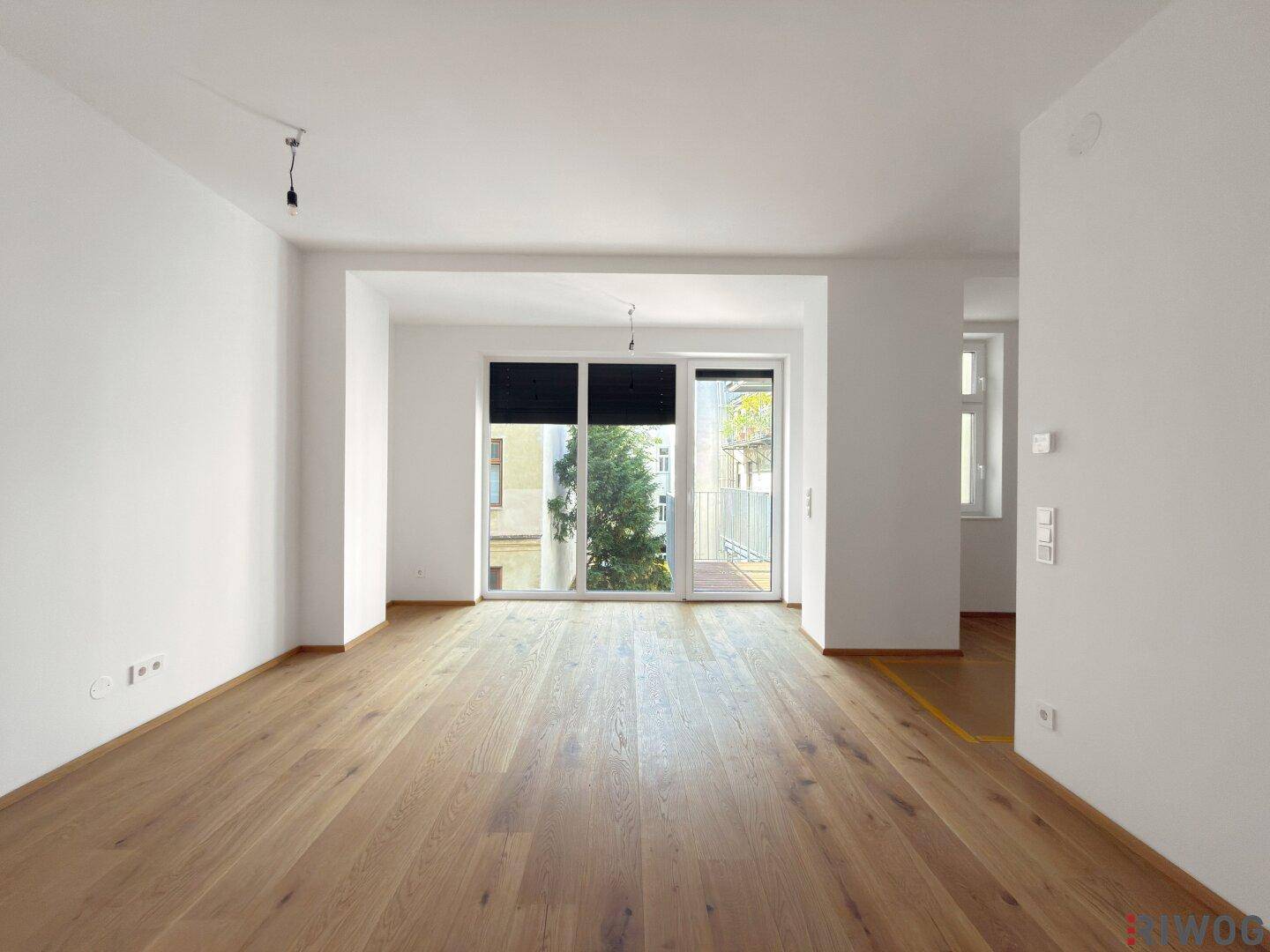 Geräumiges Wohnzimmer mit hellem Parkettboden und großen Fenstern zum Balkon.
