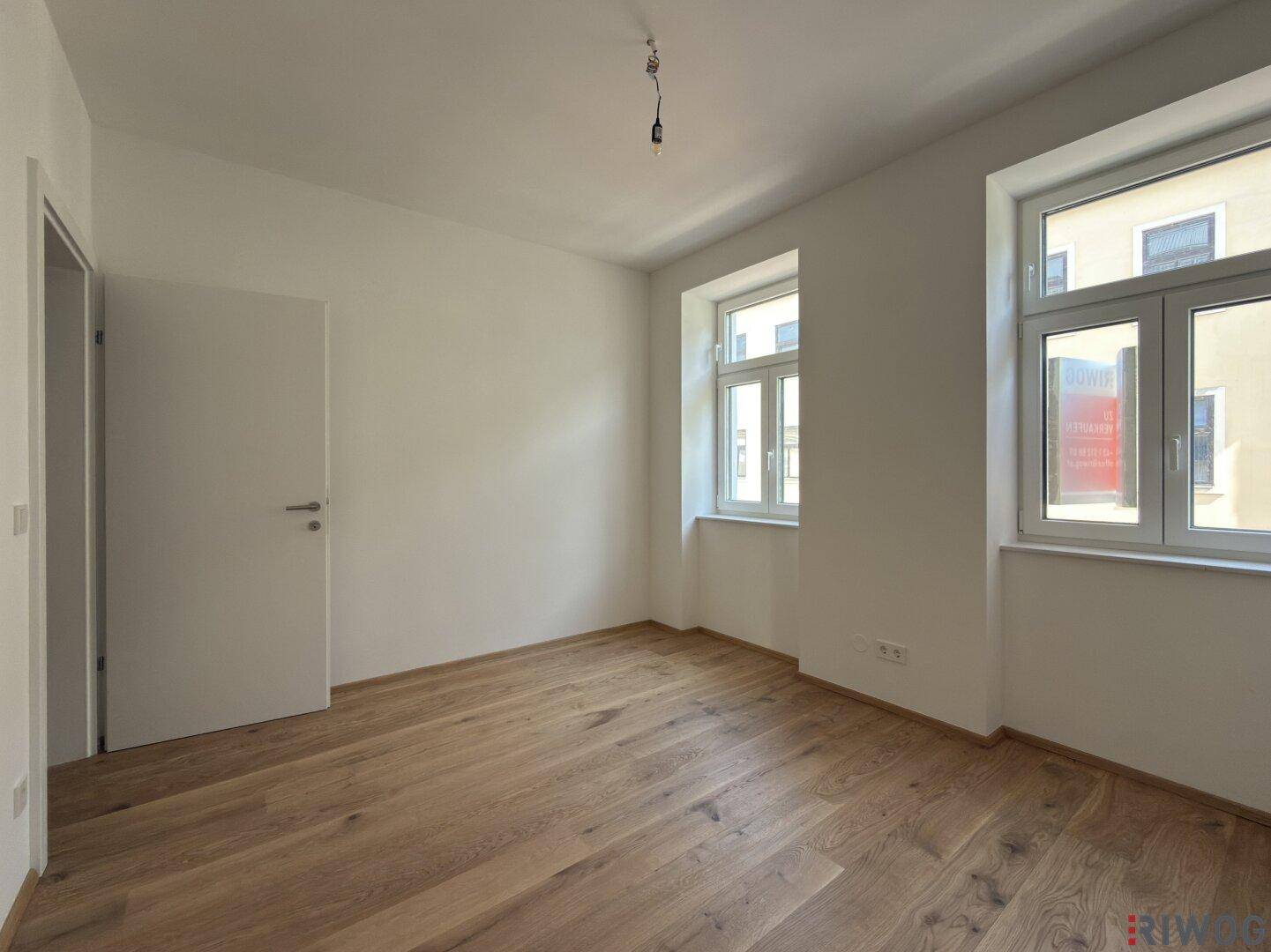 Geräumiges Schlafzimmer mit Parkettboden, zwei Fenstern und einer weißen Tür.