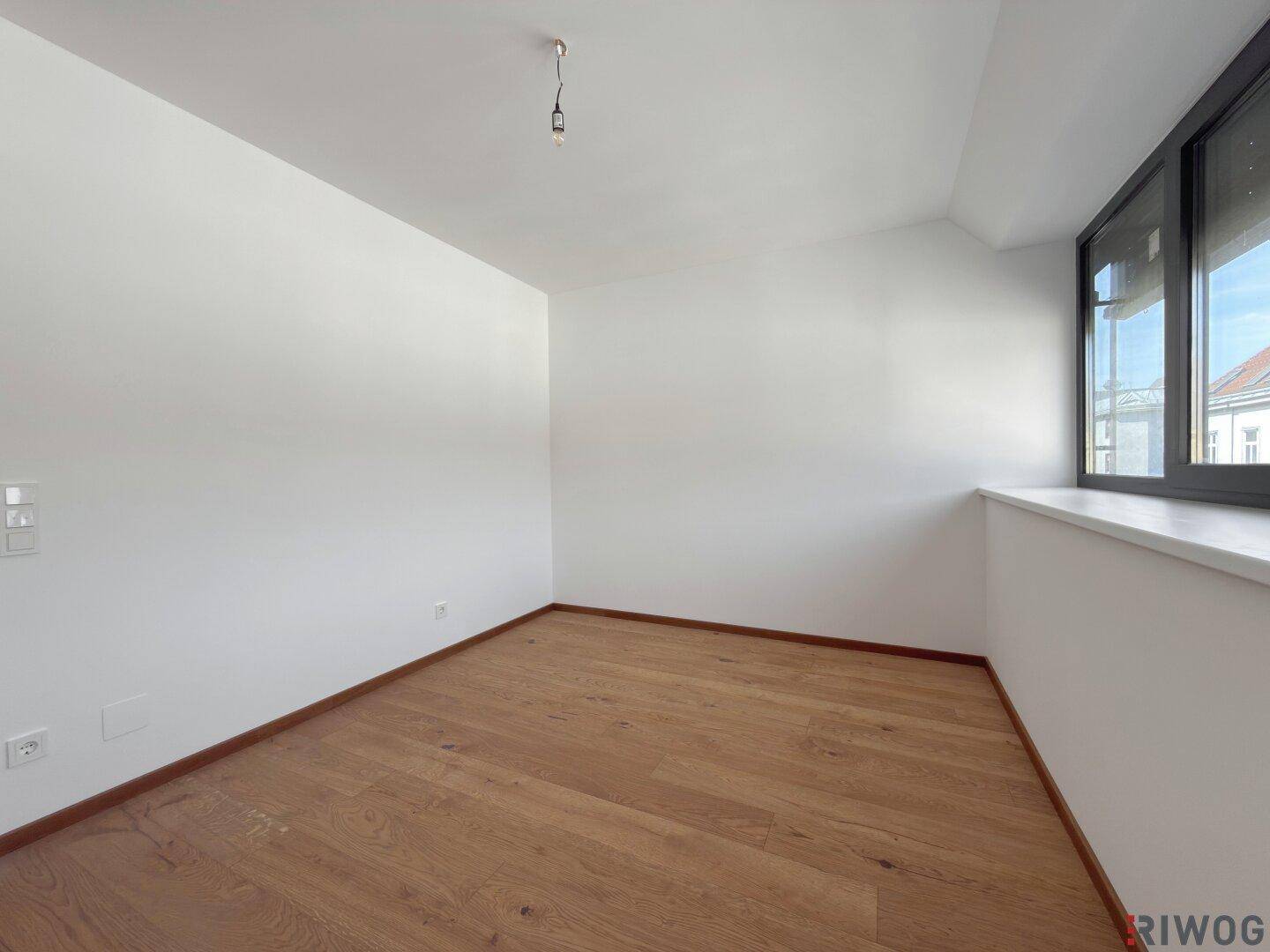 Schlichtes Zimmer mit Holzboden und einem Fenster, das für ausreichend Helligkeit sorgt.