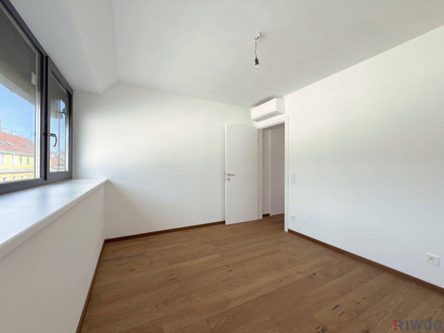 Helles Zimmer mit Holzboden, weißen Wänden und einer Klimaanlage über dem Türrahmen.