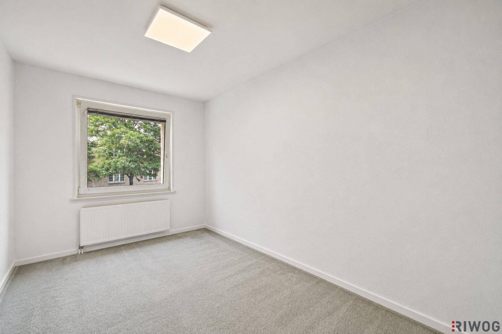 Leerer Raum mit grauem Teppichboden, einem Fenster und einem Heizkörper darunter.