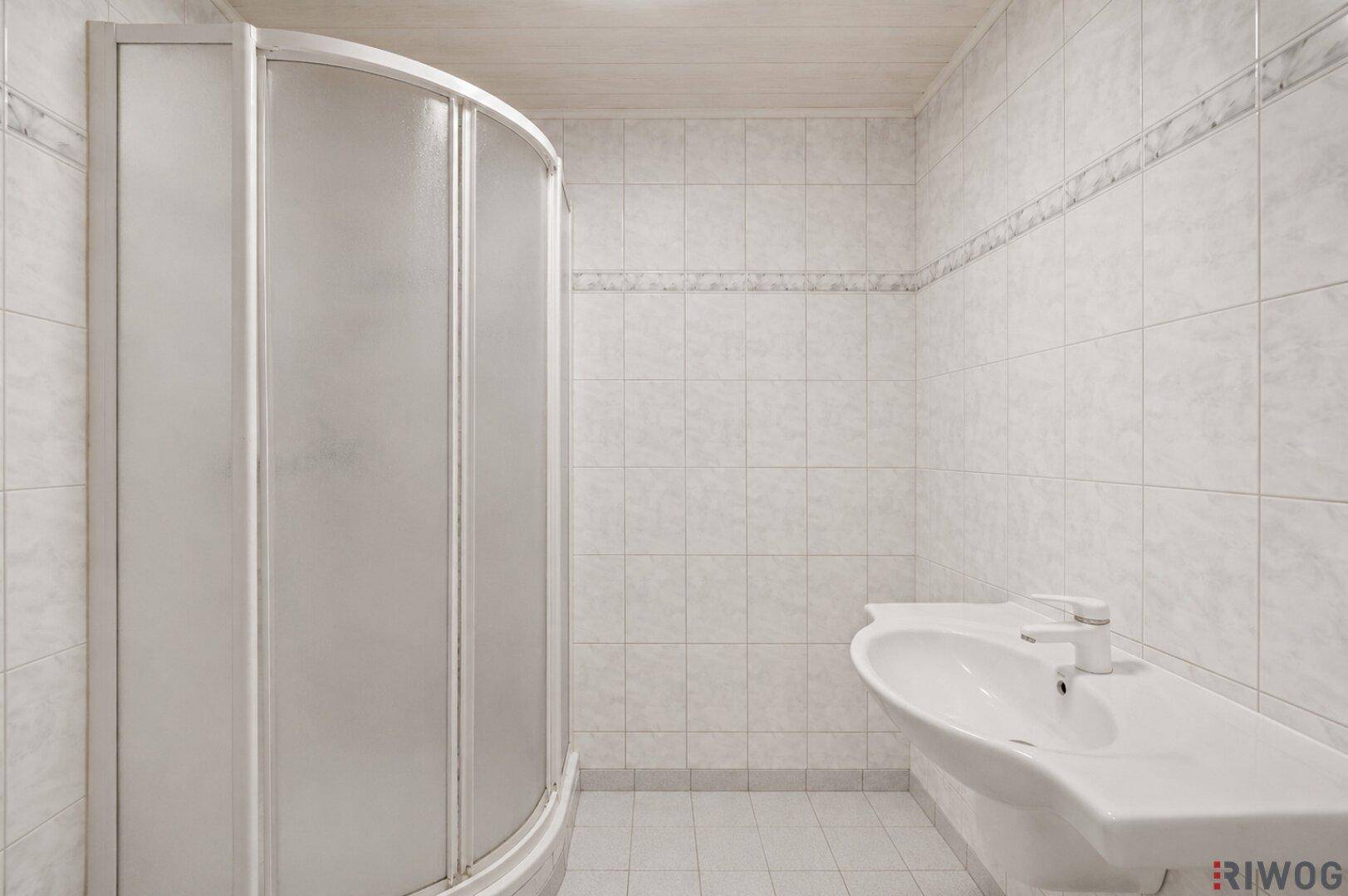 Badezimmer mit weiß gefliesten Wänden, einer Duschkabine und einem einzelnen Waschbecken.