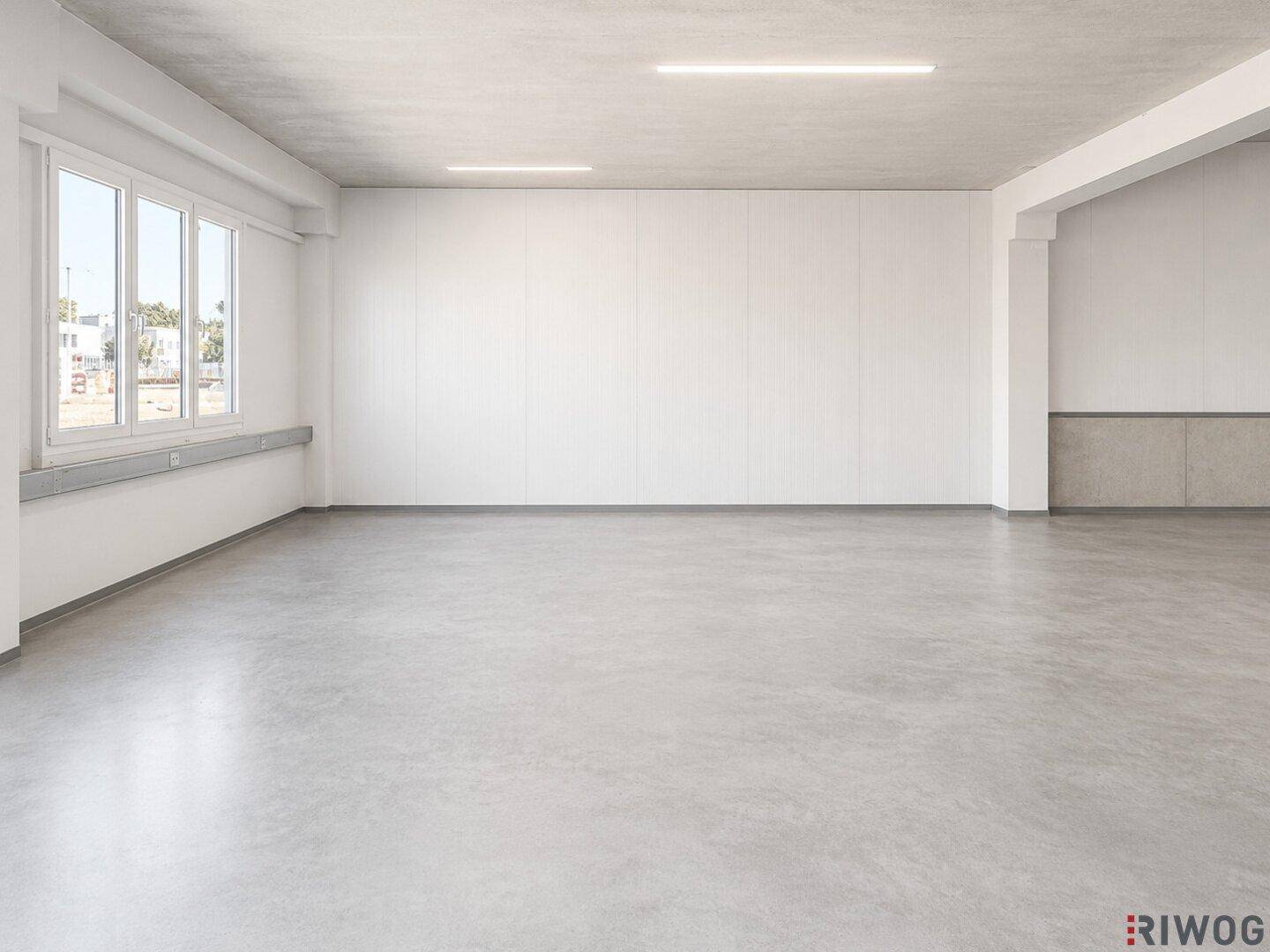 Heller, leerer Büroraum mit Betonboden, weißen Wänden und großen Fenstern für viel Tageslicht.
