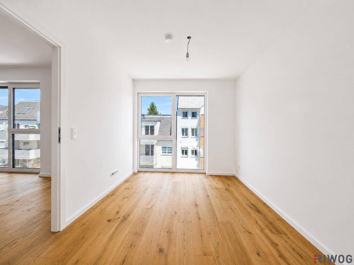 Helles Zimmer mit Holzboden und Fensterfront, die viel Tageslicht in den Raum bringt und Ausblick bietet.