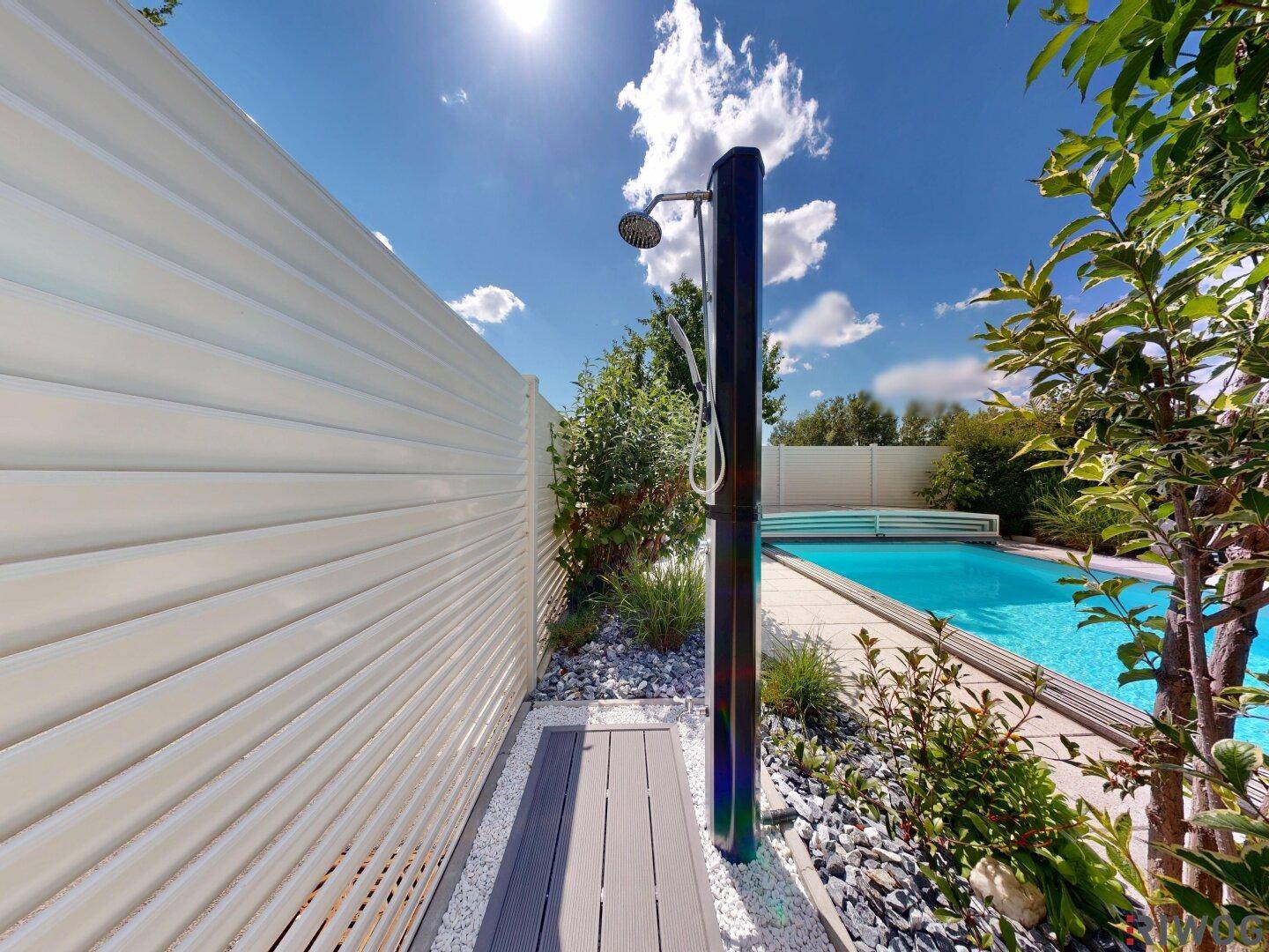 Moderne Außendusche am Poolbereich mit Kiesbeet und weißem Sichtschutz unter blauem Himmel.