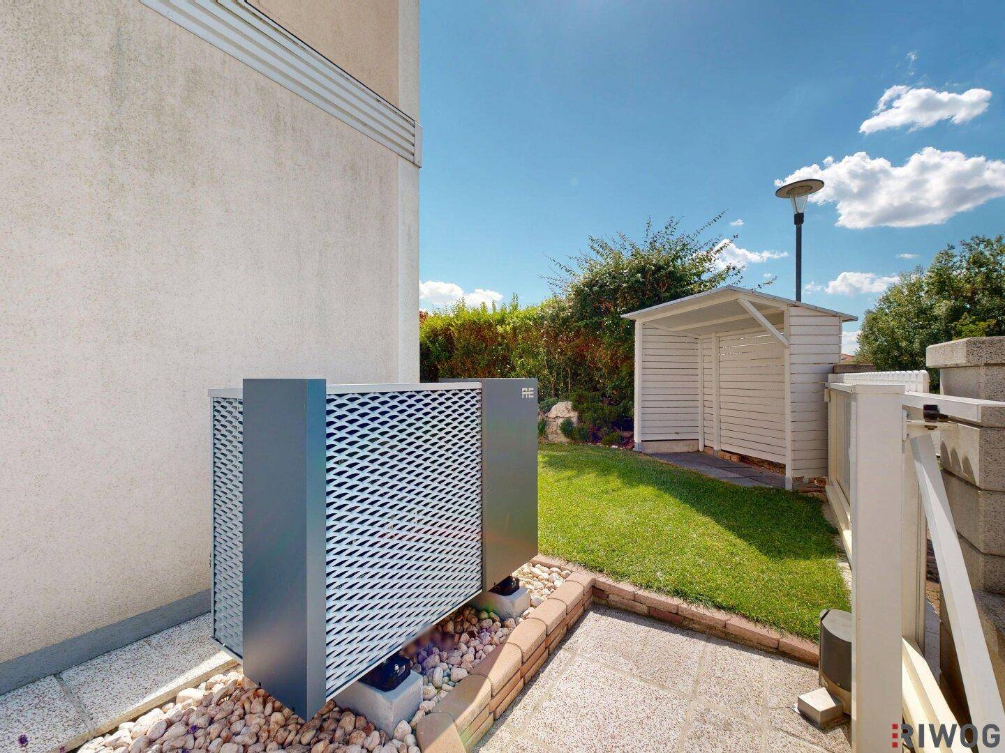 Moderne Wärmepumpe neben dem Haus im Gartenbereich mit Gartenschuppen im Hintergrund.