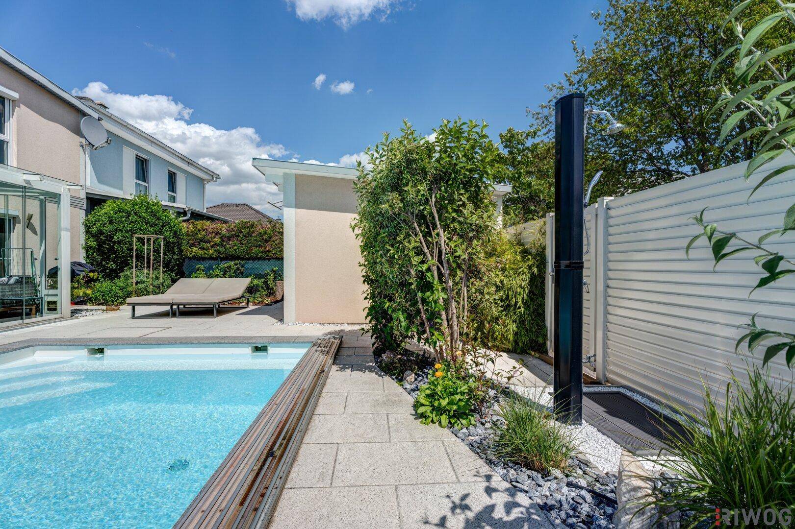 Blauer Swimmingpool mit Außendusche und gepflegter Bepflanzung im Gartenbereich.