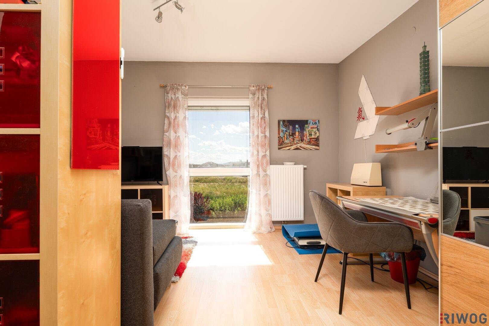 Freundliches Zimmer mit Schreibtisch, Holzboden und großem Fenster mit Ausblick.