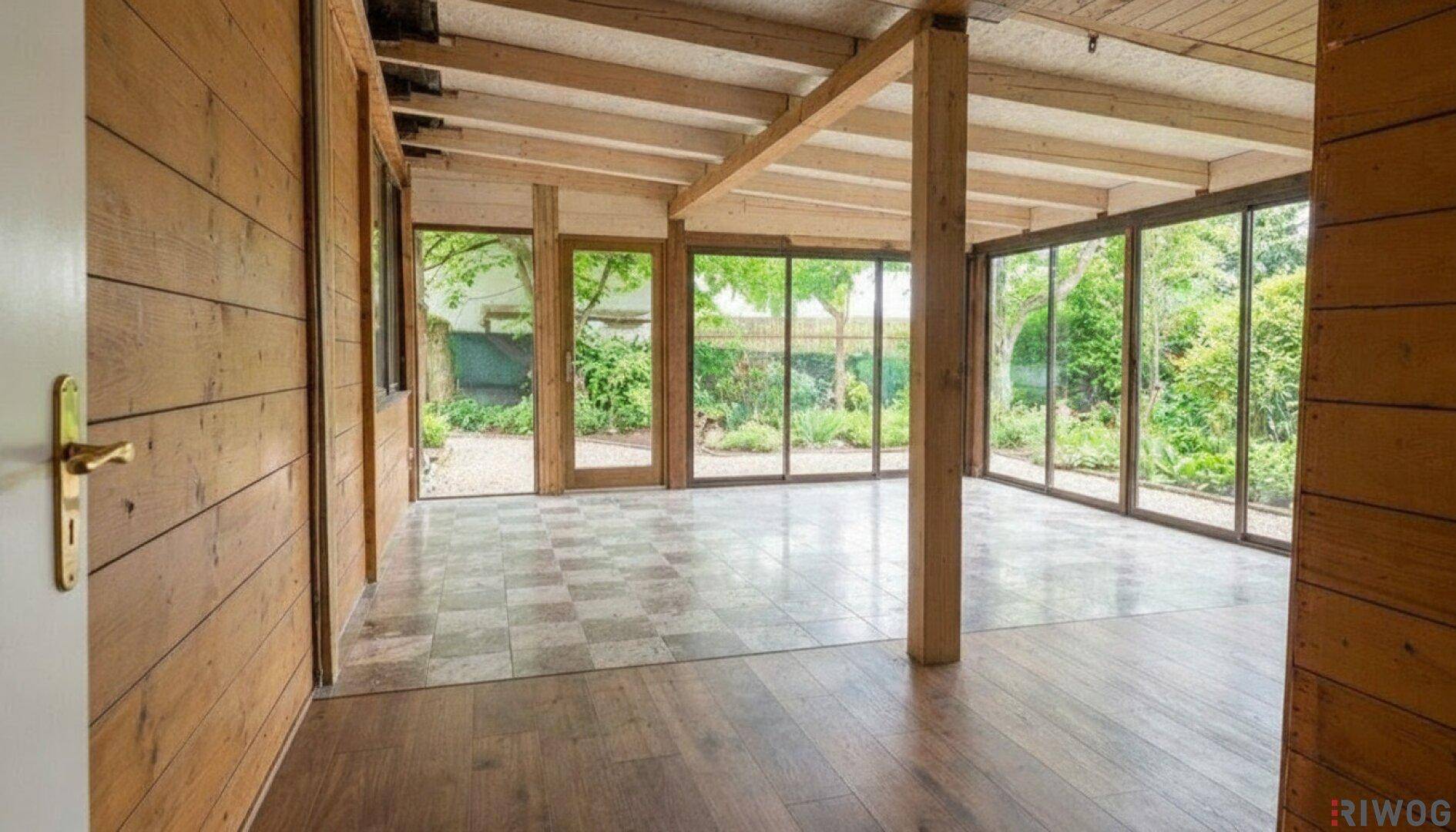 Helles Zimmer mit Holzwänden, Fliesen- und Holzboden, umgeben von großen Fenstern mit Gartenblick.